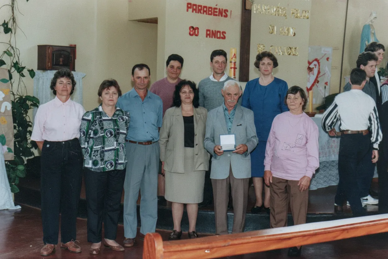 Um grupo de treze pessoas, algumas sorrindo, posa para uma foto em um evento formal, com um homem idoso no centro segurando uma pequena caixa ou placa, e uma mulher mais jovem à sua esquerda.