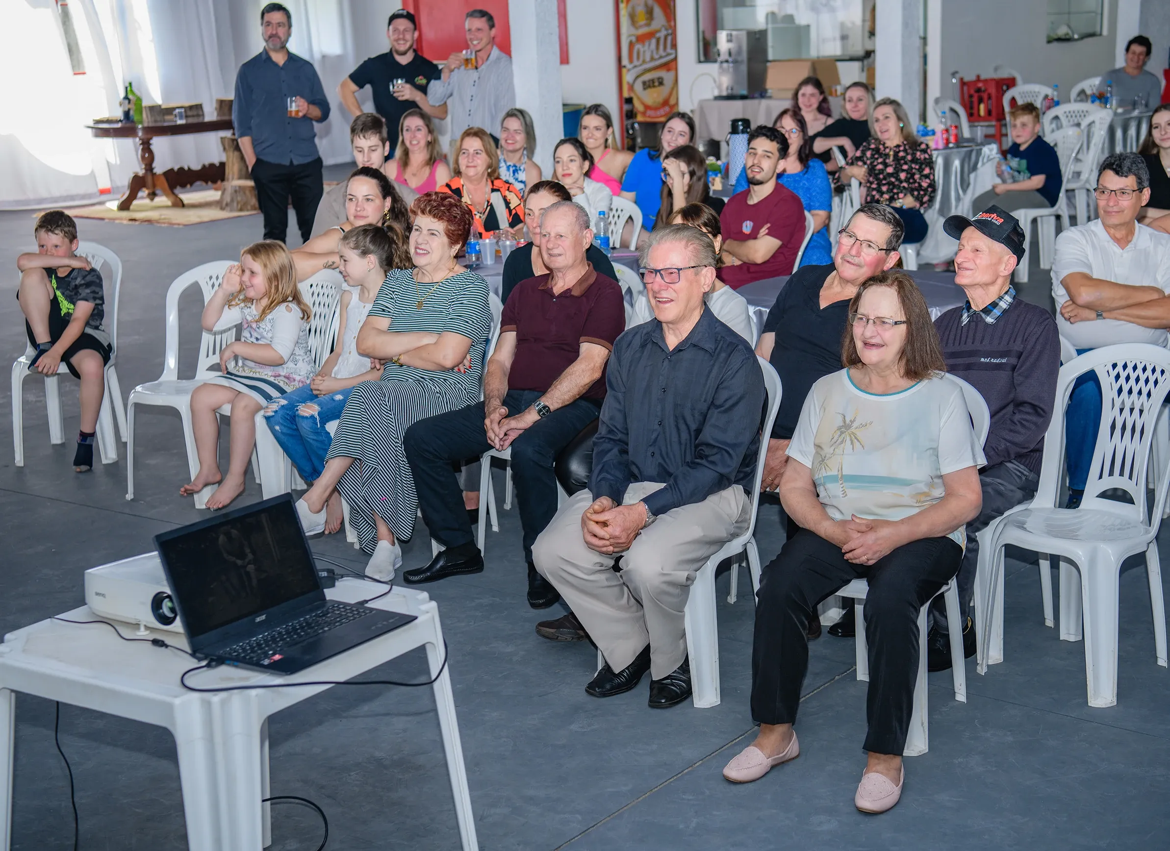 Um grupo de pessoas de diferentes idades está sentado em cadeiras de plástico brancas, olhando para a frente, onde um laptop e um projetor estão em uma mesa branca. Alguns adultos estão de pé ao fundo, e todos estão em um ambiente interno com piso cinza e paredes brancas.