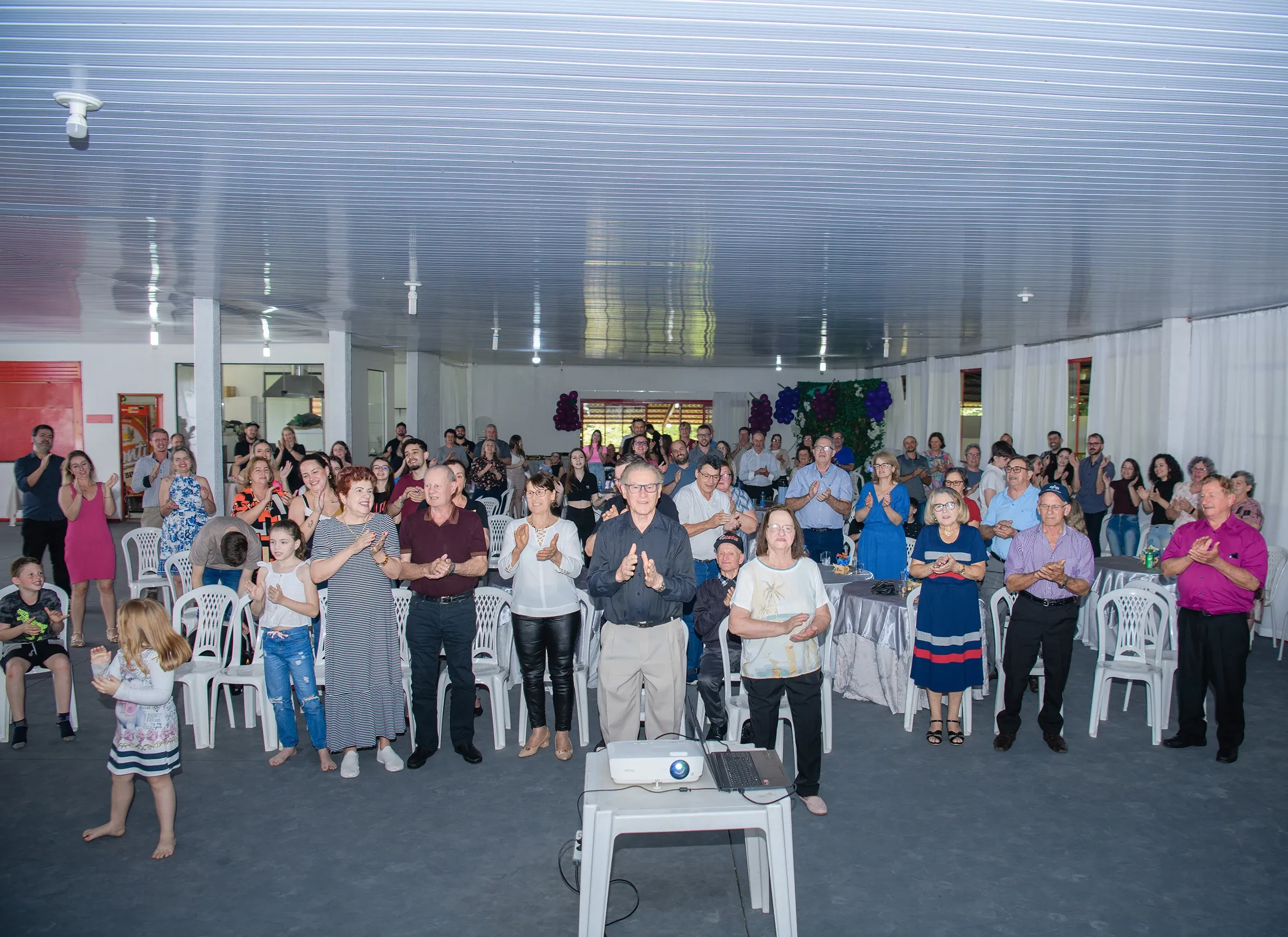 Um grupo de pessoas aplaudindo em um evento interno, com uma criança dançando na frente.