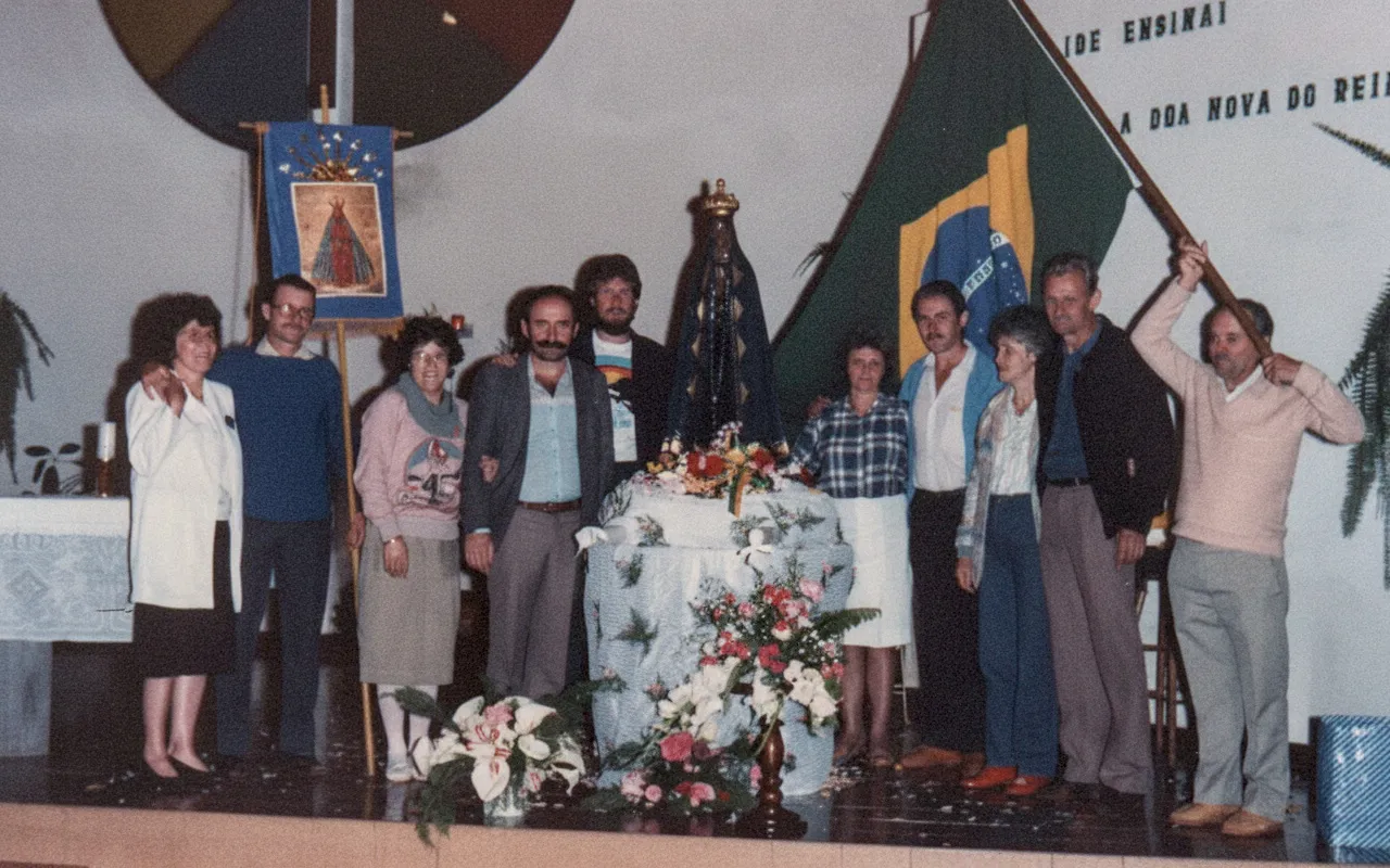 Um grupo de doze pessoas, algumas sorrindo, posa para uma foto em um palco, com alguns artefatos religiosos e uma bandeira do Brasil.