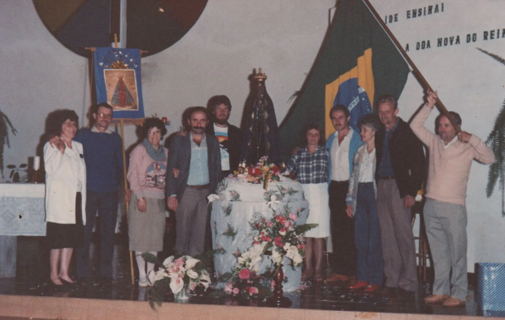 Um grupo de doze pessoas, algumas sorrindo, posa para uma foto em um palco, com alguns artefatos religiosos e uma bandeira do Brasil. (Original)