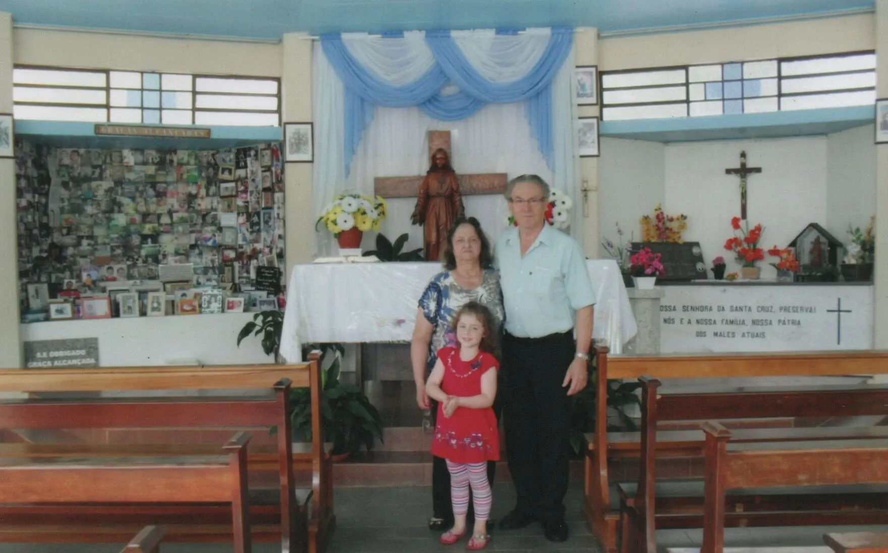 Família de três pessoas, avós e neta, em pé na frente do altar de uma capela simples.