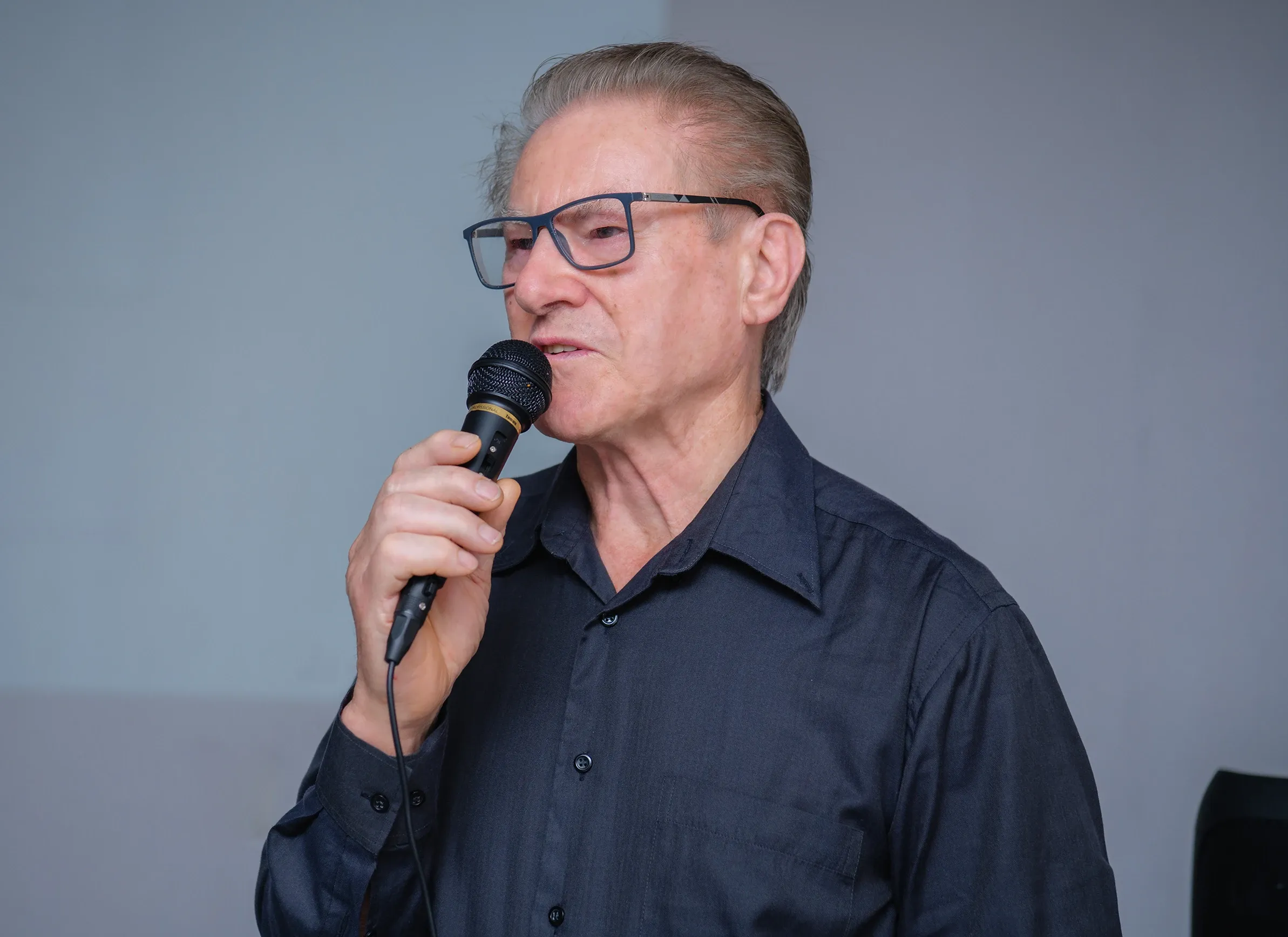 Homem idoso com óculos e camisa escura segurando um microfone, falando ou cantando.