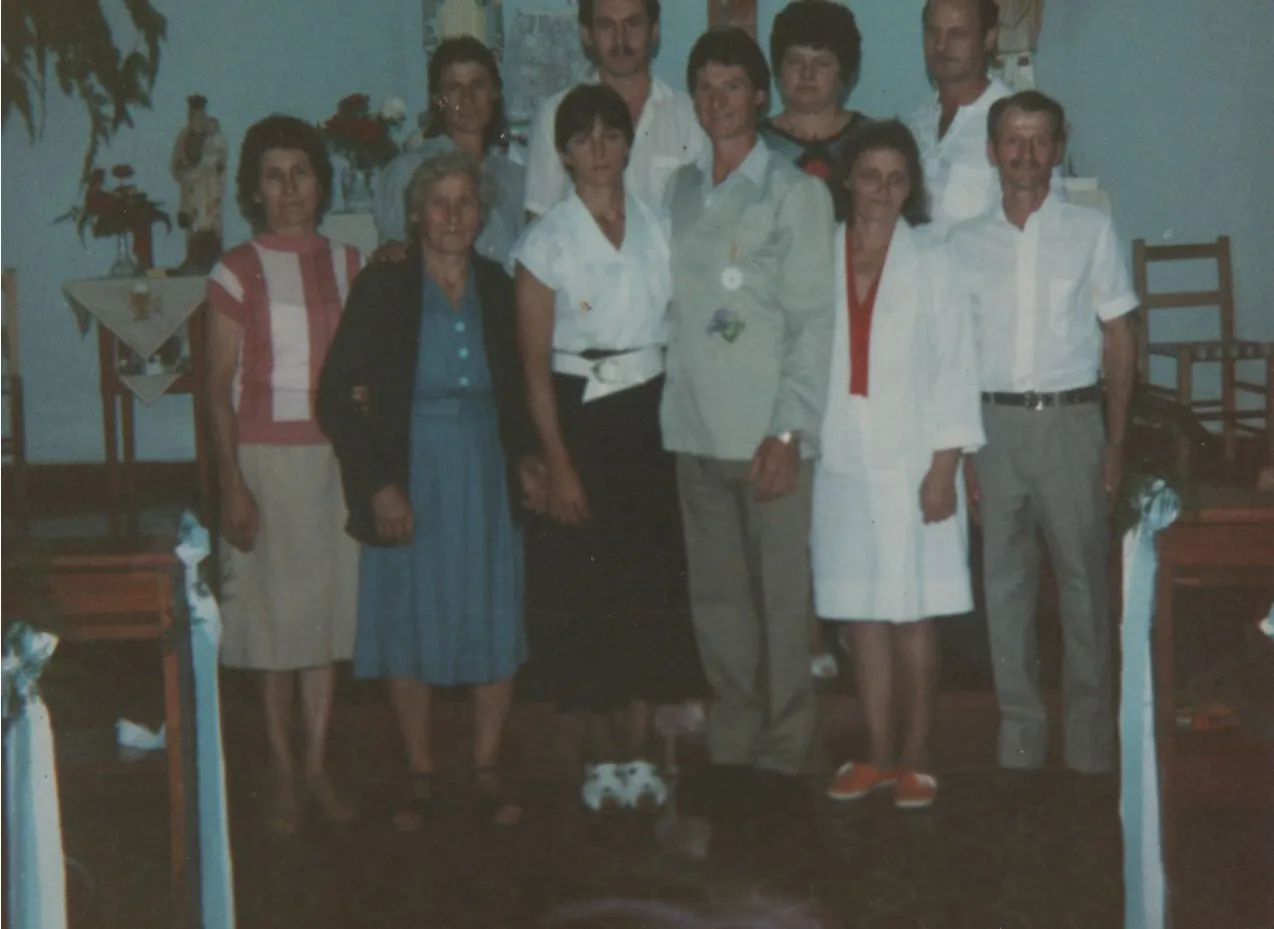 Uma fotografia envelhecida de um grande grupo de nove adultos, com seis mulheres e três homens, posando juntos num salão de reuniões com decoração de estilo igreja. (Original)