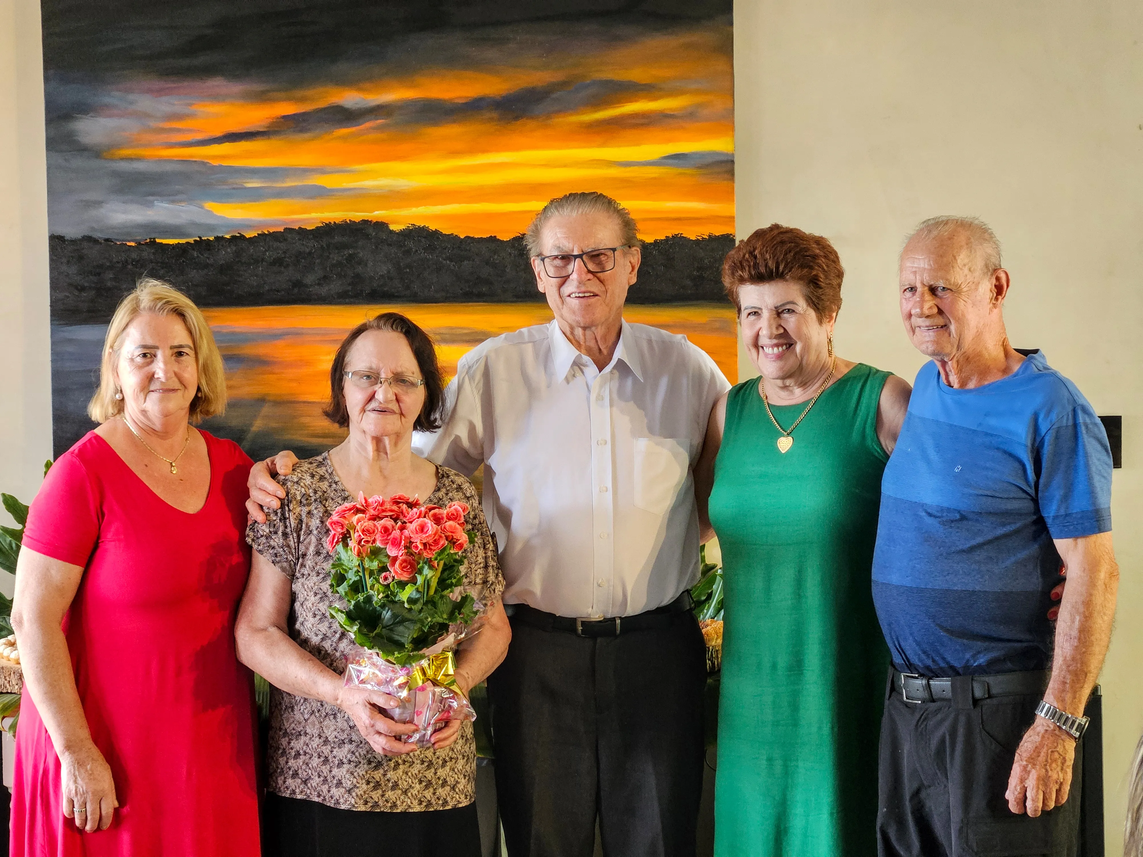 Um grupo de cinco pessoas mais velhas, quatro mulheres e um homem, posa para uma foto na frente de uma pintura de pôr do sol, enquanto uma das mulheres segura um buquê de flores.