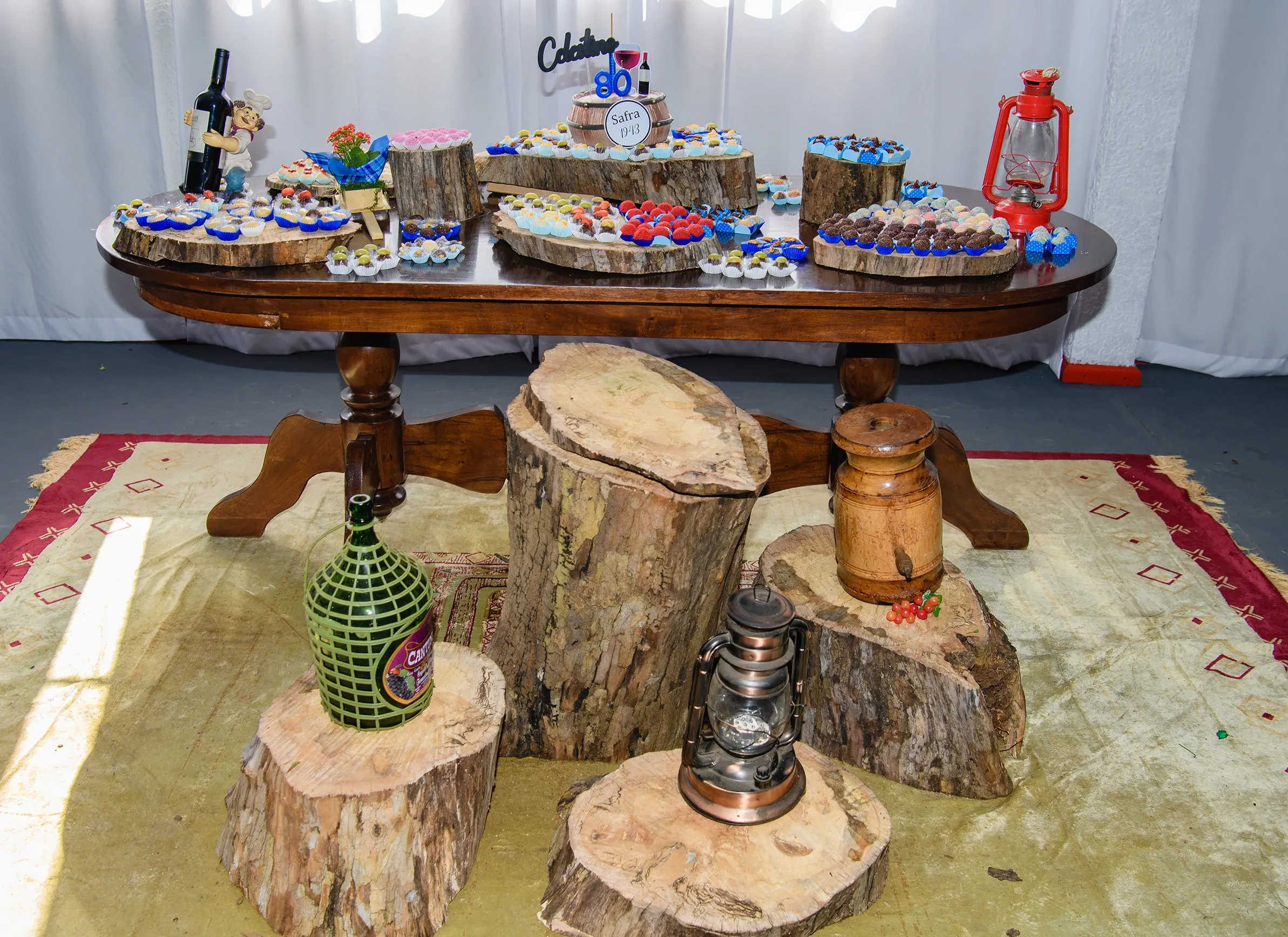 Uma mesa de madeira rústica com vários bolos pequenos e doces dispostos em fatias de tronco de árvore, decorada com uma garrafa de vinho, uma lamparina vermelha e outra marrom, sobre um tapete amarelo com bordas vermelhas.