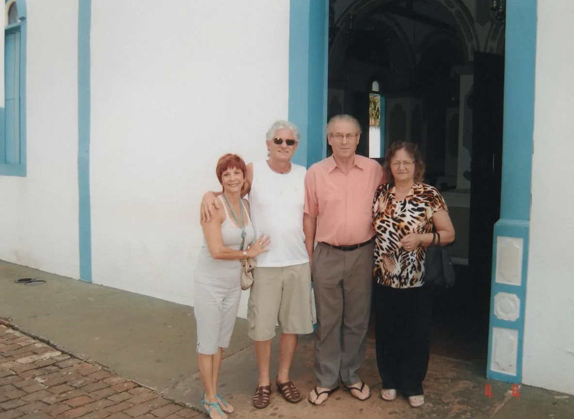 Quatro adultos - duas mulheres e dois homens - de pé na frente de uma igreja caiada de branco com acabamentos azuis, sorrindo para a câmera.