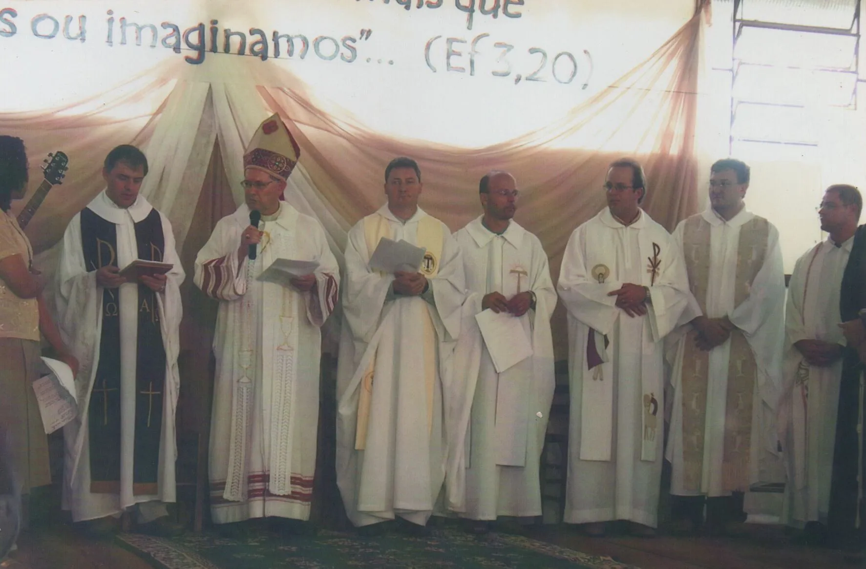 Vários bispos e sacerdotes em vestes clericais participam de um evento religioso sob uma faixa com texto em português. (Original)