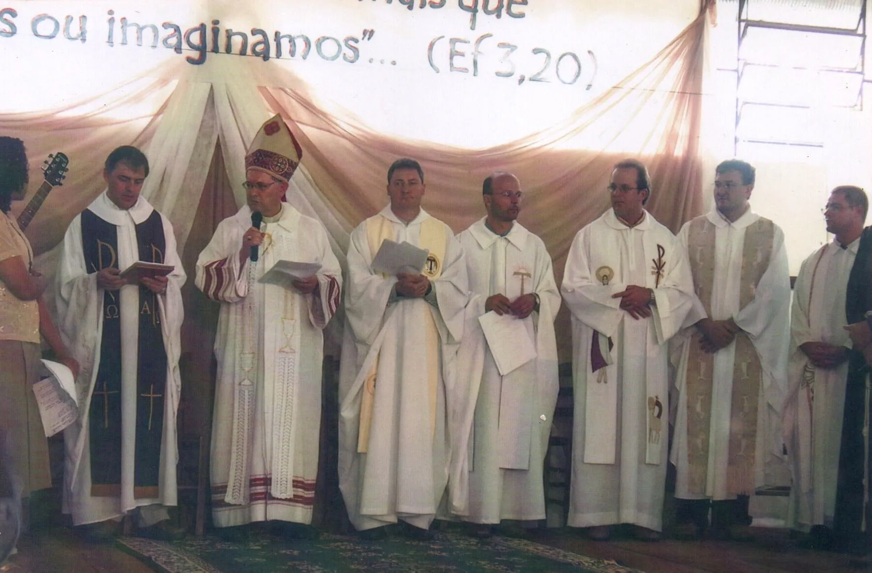 Vários bispos e sacerdotes em vestes clericais participam de um evento religioso sob uma faixa com texto em português.
