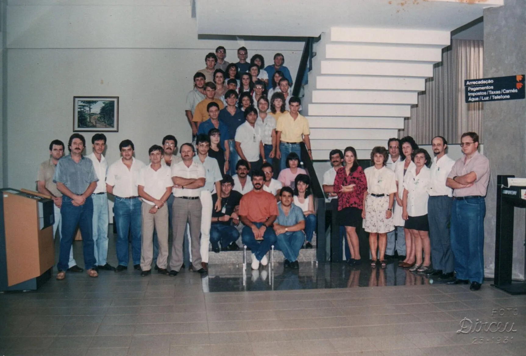 Grupo de cerca de 30 a 40 pessoas, maioritariamente homens, posa em escadas e chão interior, com alguns em pé e sentados na frente.