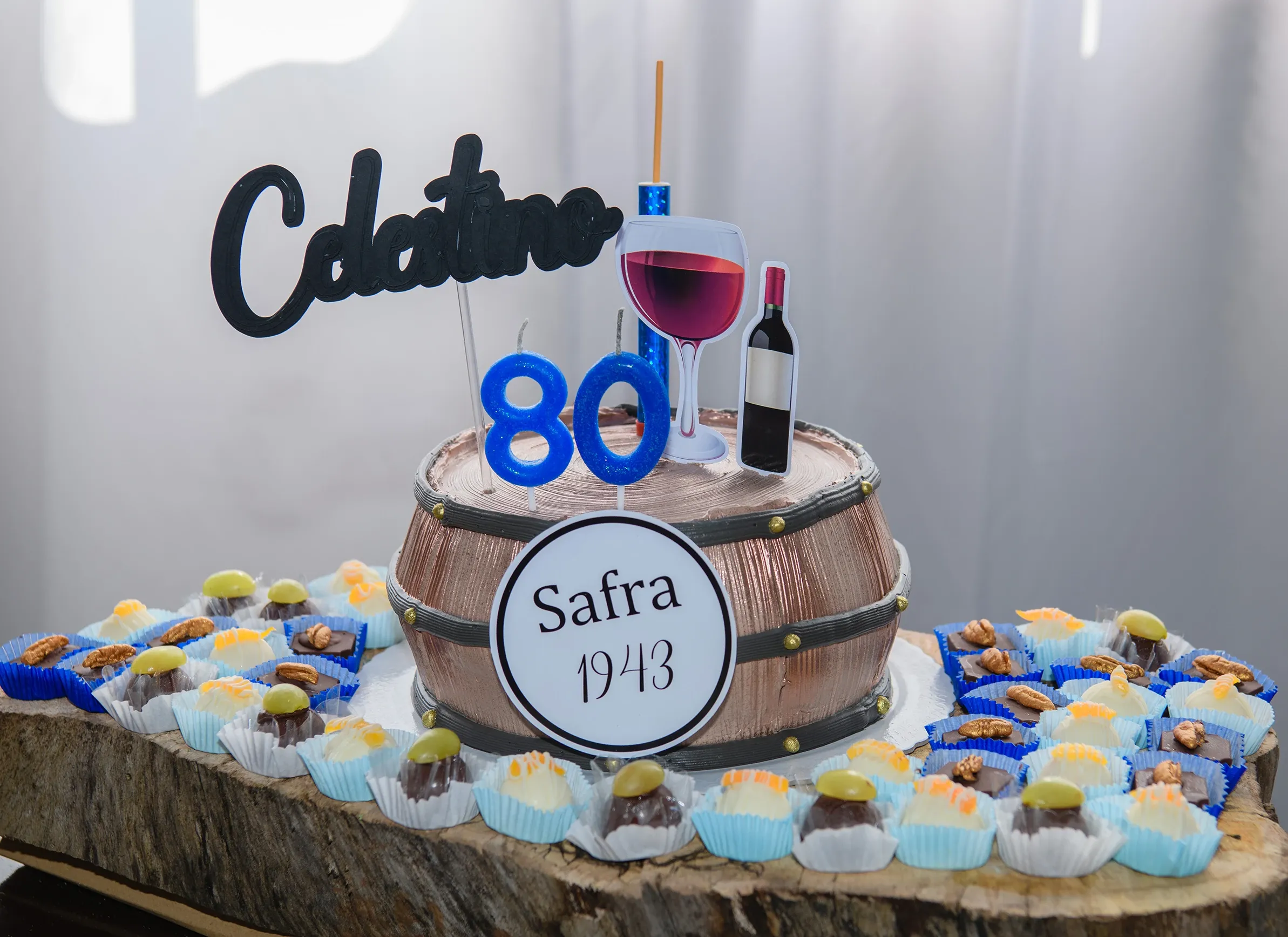 Um bolo de aniversário em formato de barril de vinho com velas de número 80, um topo de bolo com "Celestino" e "Safra 1943", rodeado por doces em uma tábua de madeira.