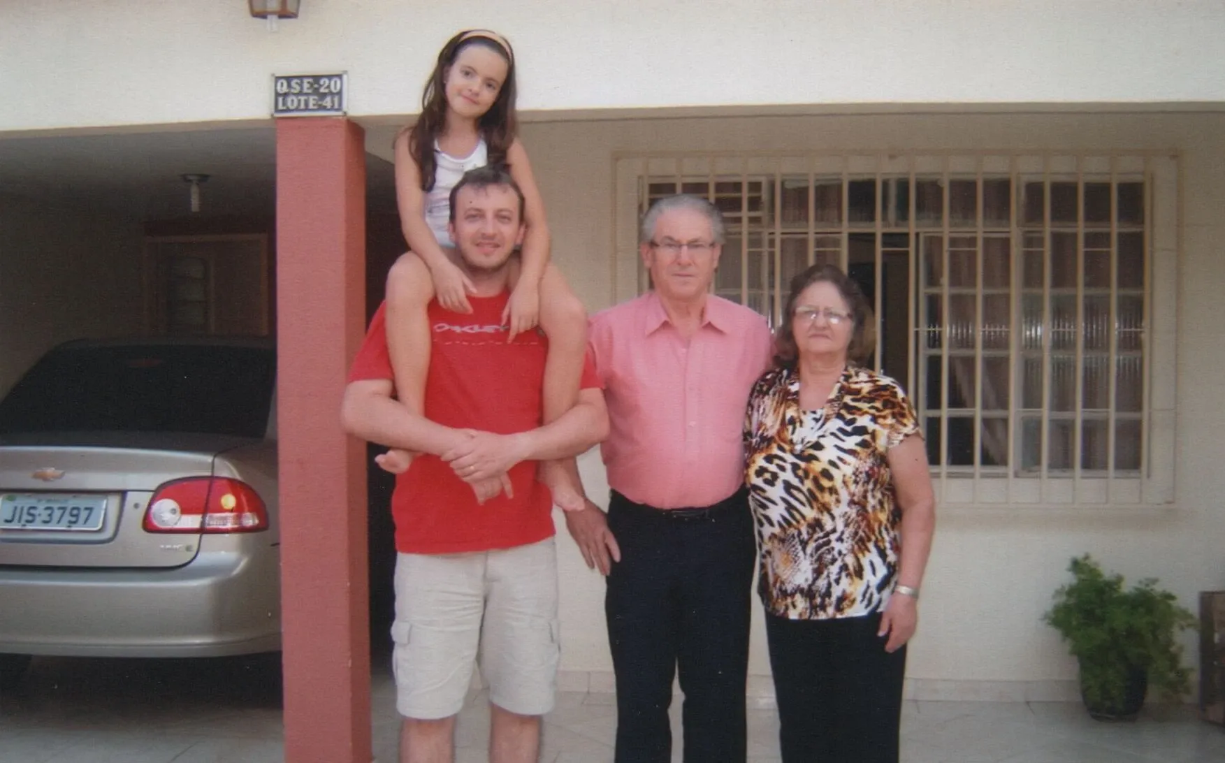 Uma jovem garota sentada nos ombros de um homem sorrindo para a câmera, enquanto uma mulher e um homem idosos estão ao lado deles, em frente a uma casa.