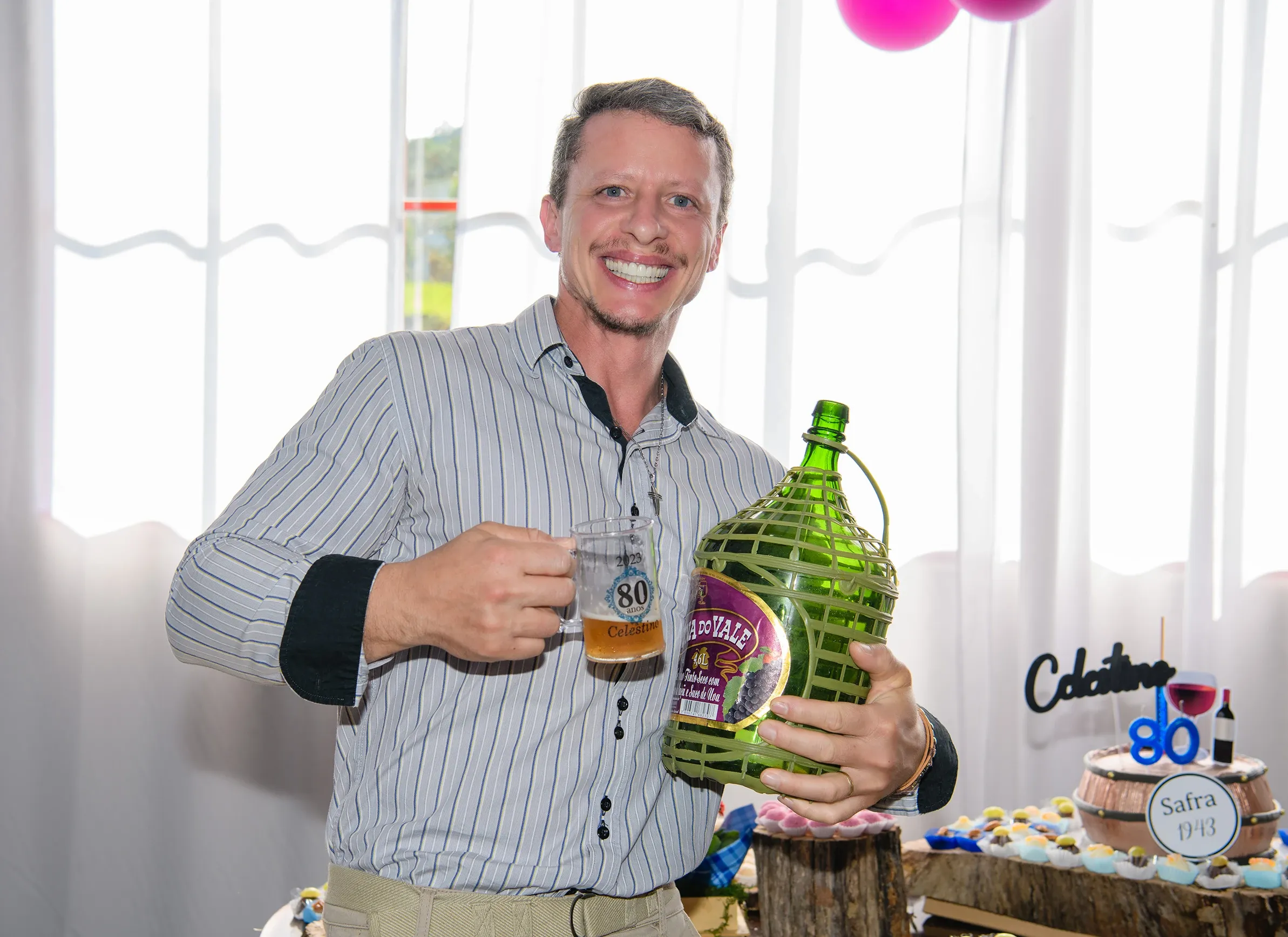 Homem de camisa listrada sorrindo e segurando uma caneca de chope "80 Celesia" e uma garrafa grande de vinho verde, com uma mesa de sobremesas ao fundo.