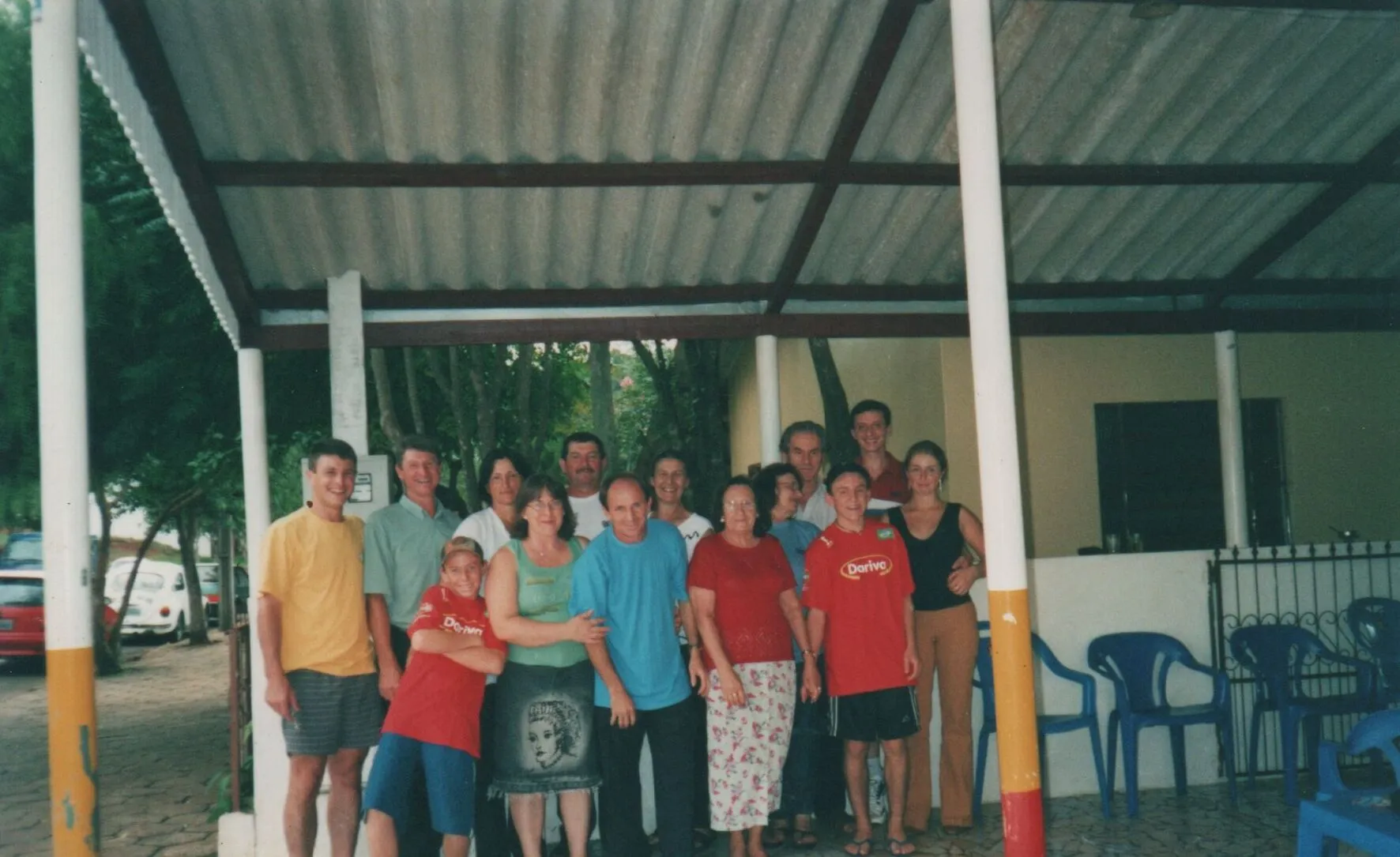 Um grupo de quinze adultos sorridentes, de várias idades e etnias, posa para uma foto sob um telhado de metal corrugado, com folhagem verde ao fundo. (Original)