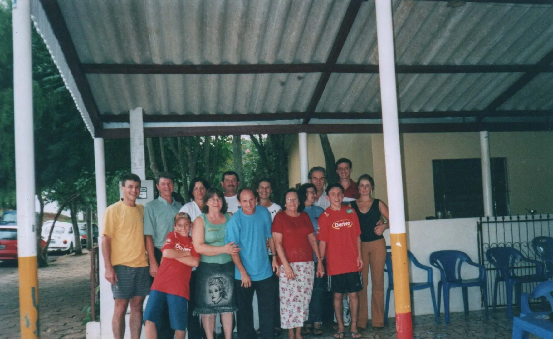 Um grupo de quinze adultos sorridentes, de várias idades e etnias, posa para uma foto sob um telhado de metal corrugado, com folhagem verde ao fundo.