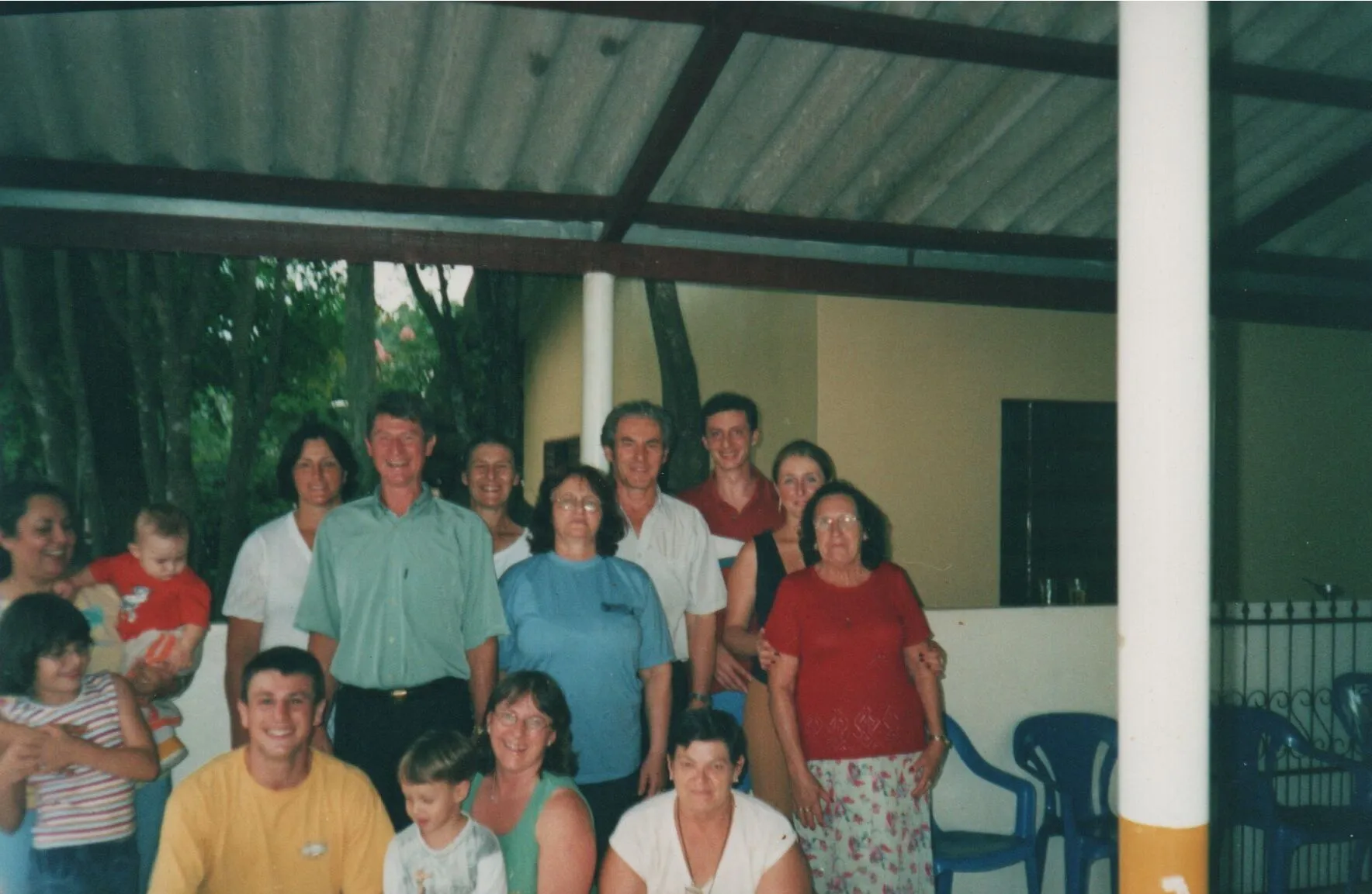 Um grupo de pessoas de diferentes idades posa para uma foto em um espaço ao ar livre com um telhado e pilares, com folhagem verde ao fundo. (Original)