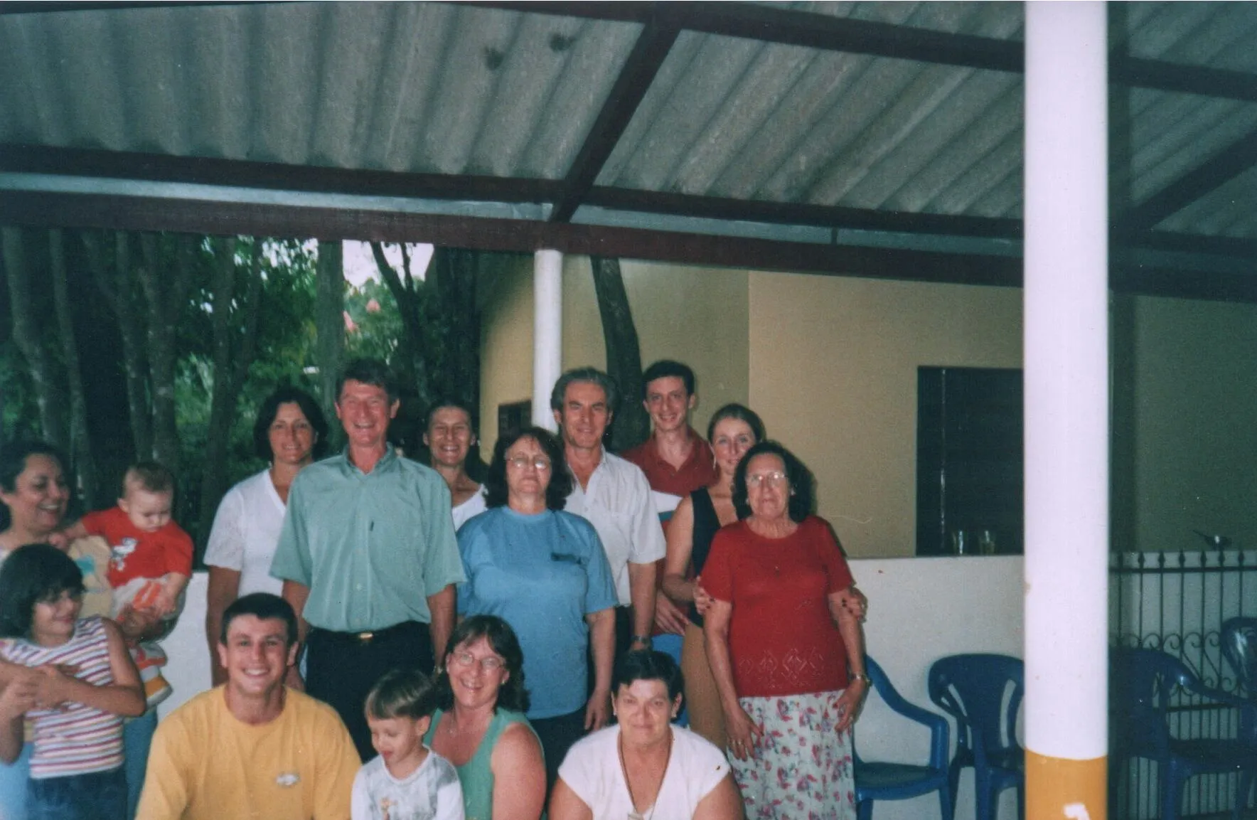 Um grupo de pessoas de diferentes idades posa para uma foto em um espaço ao ar livre com um telhado e pilares, com folhagem verde ao fundo.
