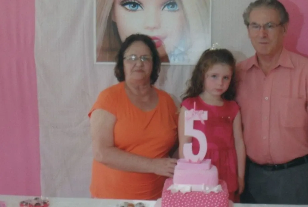 Uma menina de 5 anos de cabelo encaracolado e um vestido rosa está ao lado de seus avós em sua festa de aniversário.