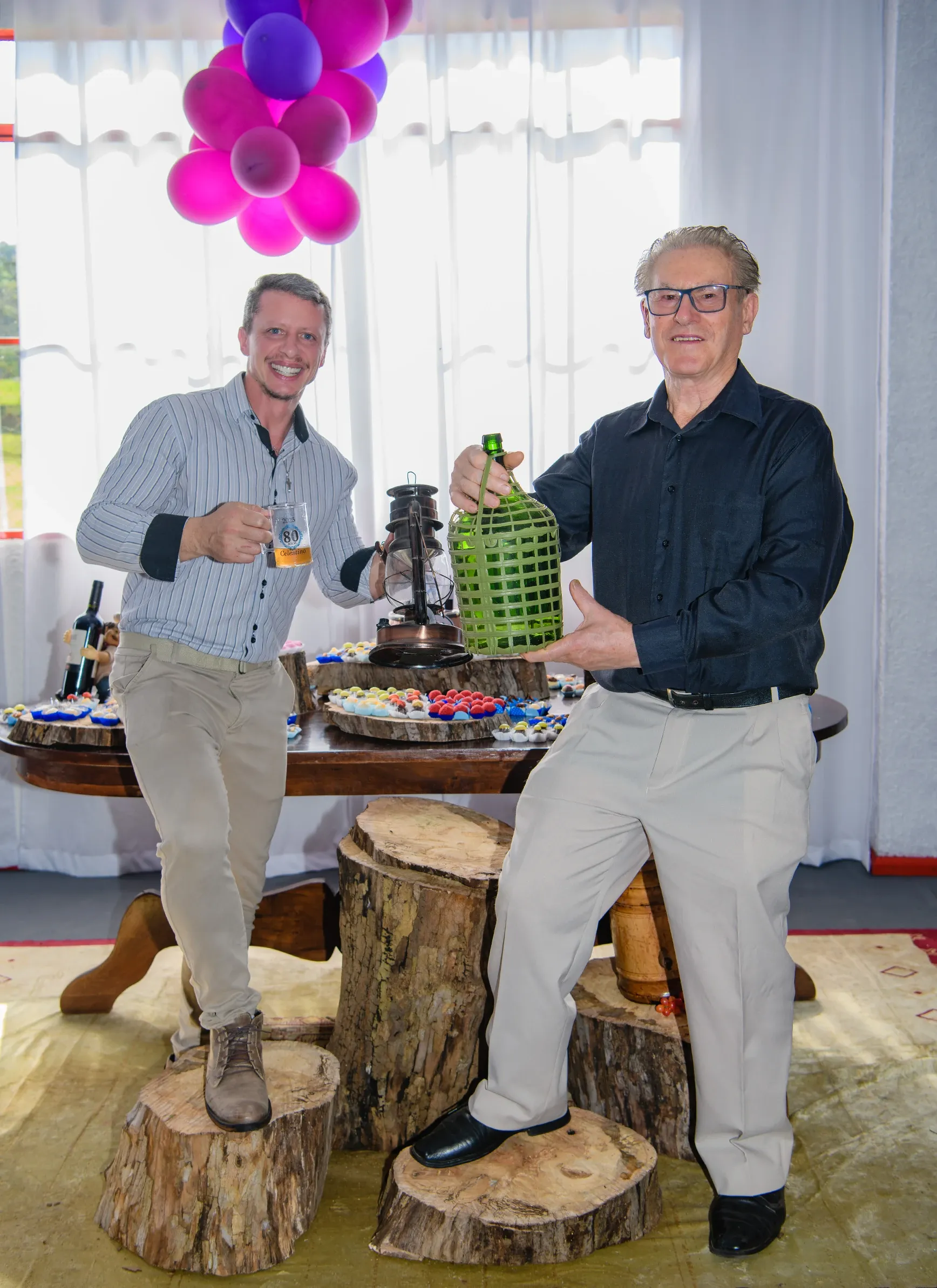 Dois homens sorridentes posam em uma festa; um segura uma caneca de cerveja e uma lanterna vintage, o outro exibe uma garrafa verde grande sobre bases de tronco de árvore, com balões roxos e rosa ao fundo.