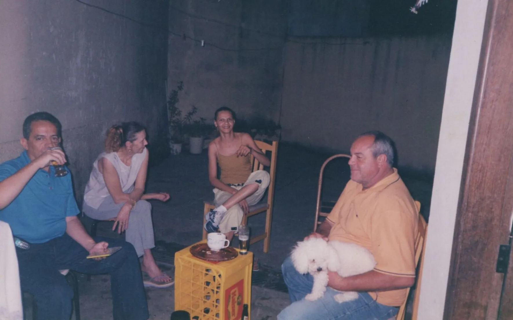 Quatro adultos sentados em um pátio mal iluminado em cadeiras de jardim, um segurando um pequeno cachorro branco e outro segurando uma cerveja. (Original)