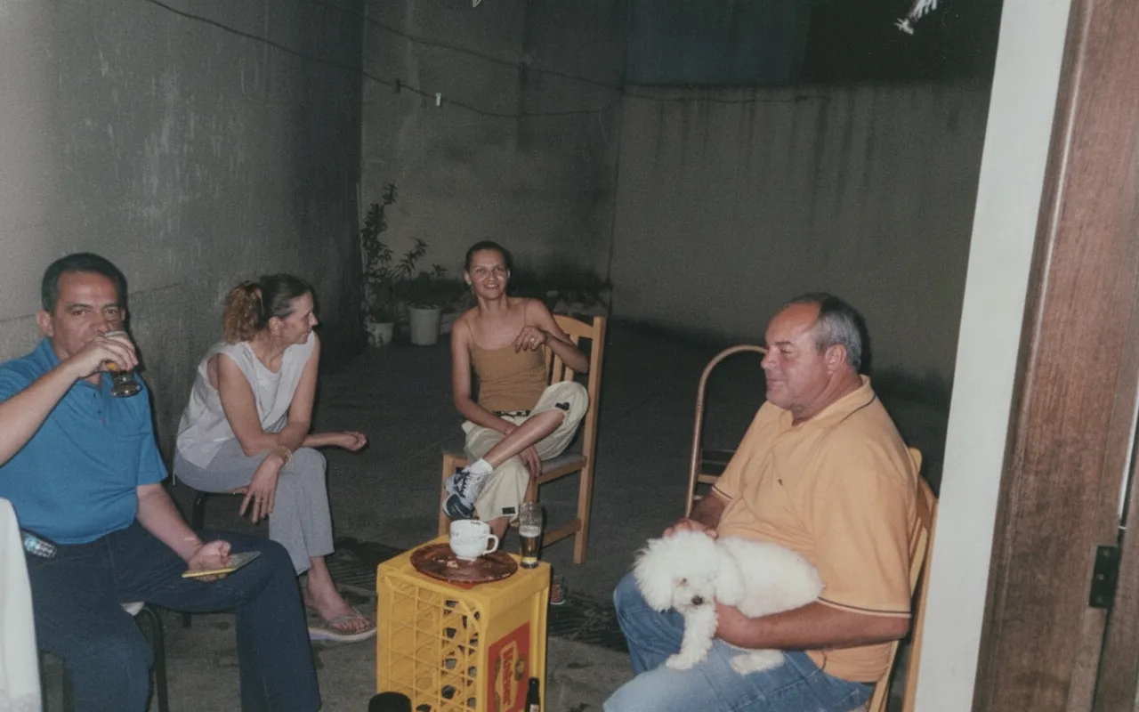Quatro adultos sentados em um pátio mal iluminado em cadeiras de jardim, um segurando um pequeno cachorro branco e outro segurando uma cerveja.
