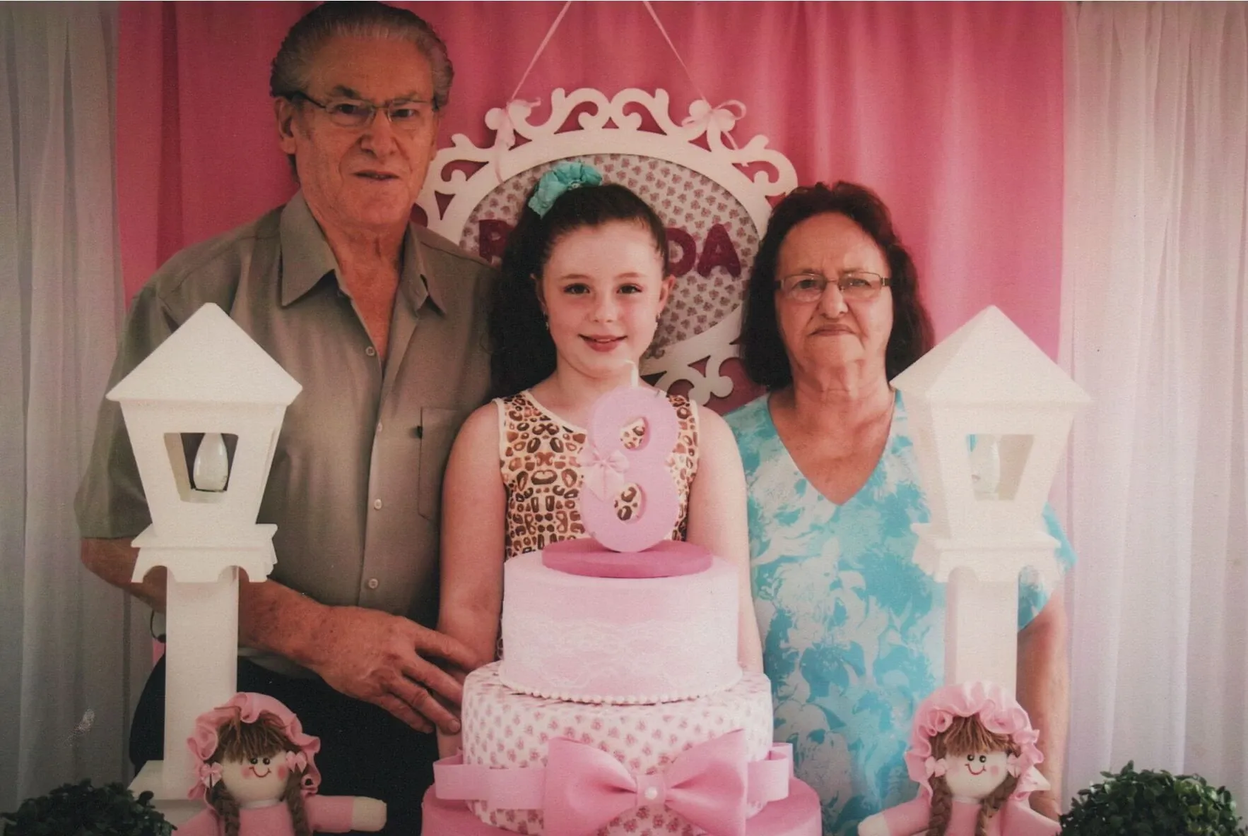 Uma menina de oito anos sorri com os avós em uma festa de aniversário com tema rosa.
