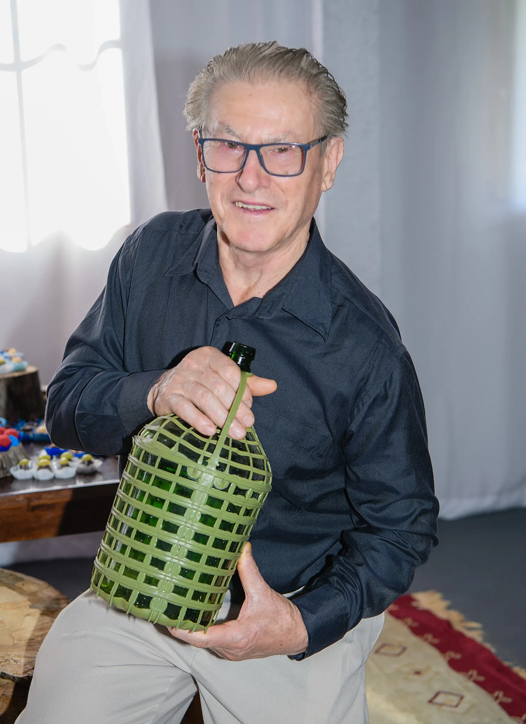 Homem mais velho, de óculos, camisa social preta e calças cáqui, segura uma garrafa de vidro verde envolta em uma cesta de vime, sorrindo para a câmera em um ambiente interno.