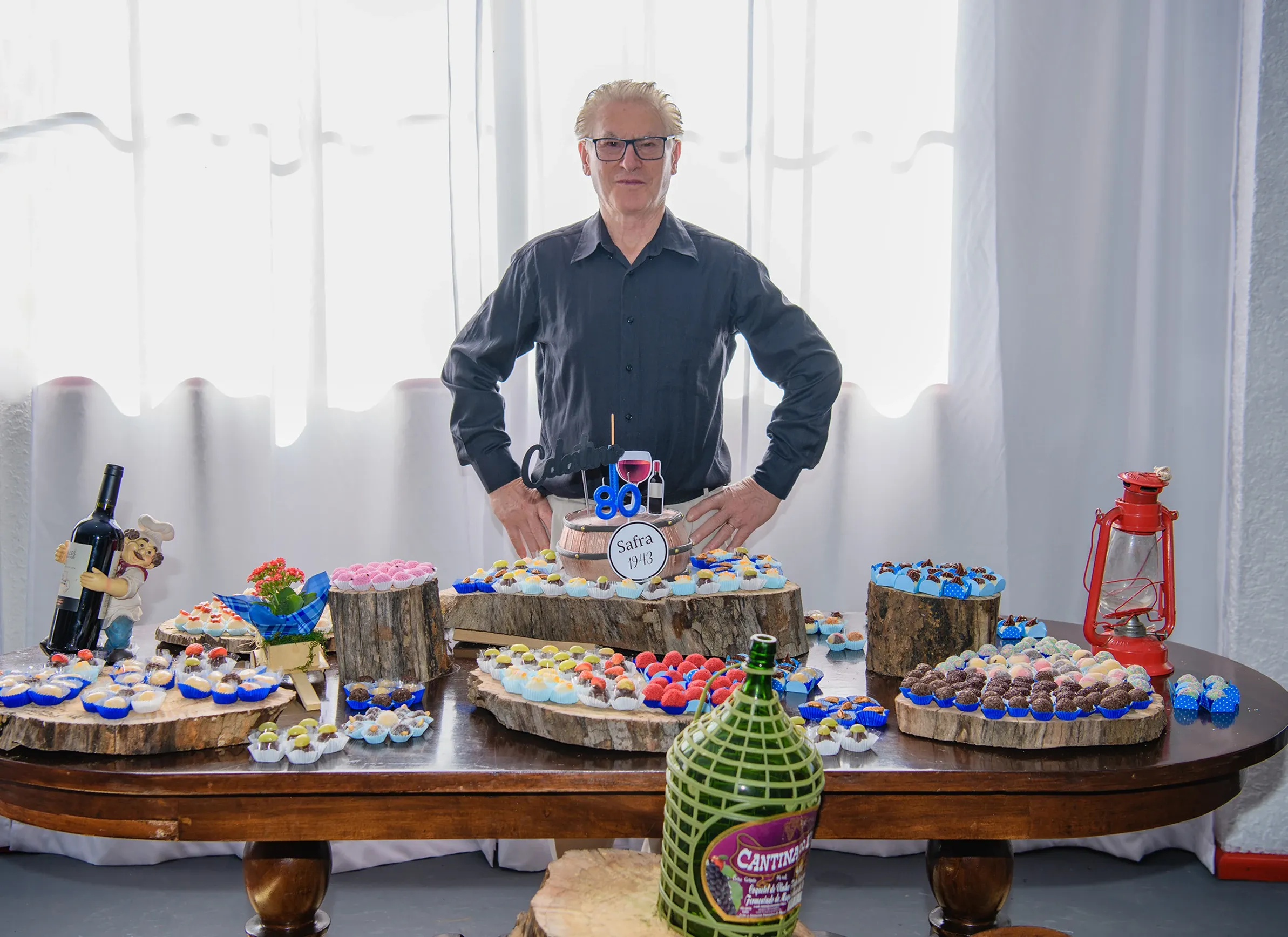 Homem de camisa preta e óculos está atrás de uma mesa de madeira decorada com vários doces em bandejas e suportes de tronco de árvore, com cortinas brancas ao fundo.