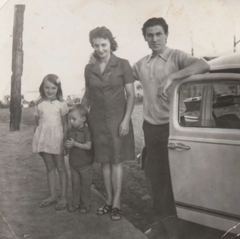 Uma família de quatro pessoas, incluindo um homem, uma mulher e duas crianças, posa ao lado de um carro azul-acinzentado, com o homem de camisa polo azul e a mulher de vestido verde, a menina de branco e o menino de camisa listrada, todos com sandálias, e um céu claro e a água à distância. (Original)