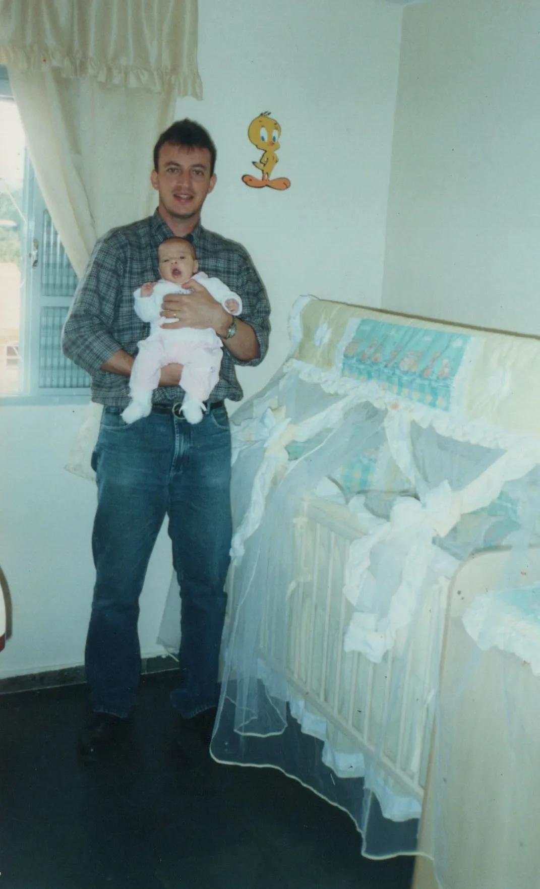 Um homem adulto em uma camisa xadrez e jeans segura um bebê com a boca aberta em um berçário com um adesivo de parede do Piu-Piu. (Original)