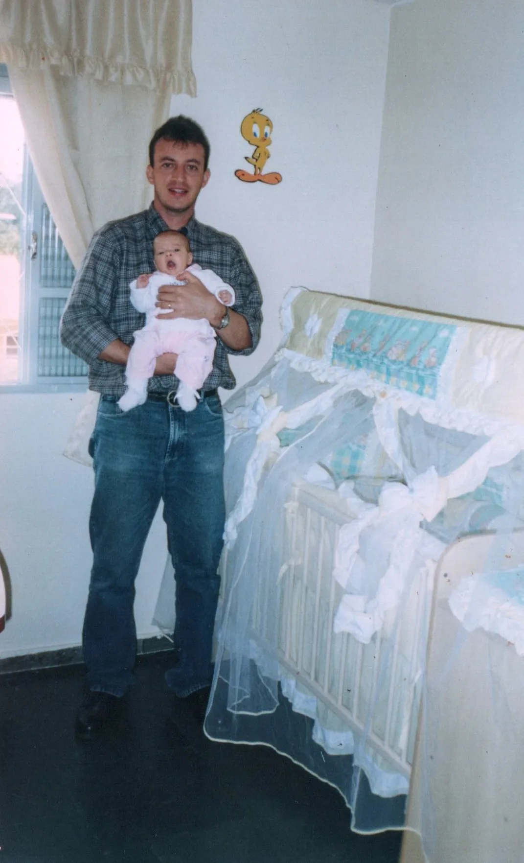 Um homem adulto em uma camisa xadrez e jeans segura um bebê com a boca aberta em um berçário com um adesivo de parede do Piu-Piu.