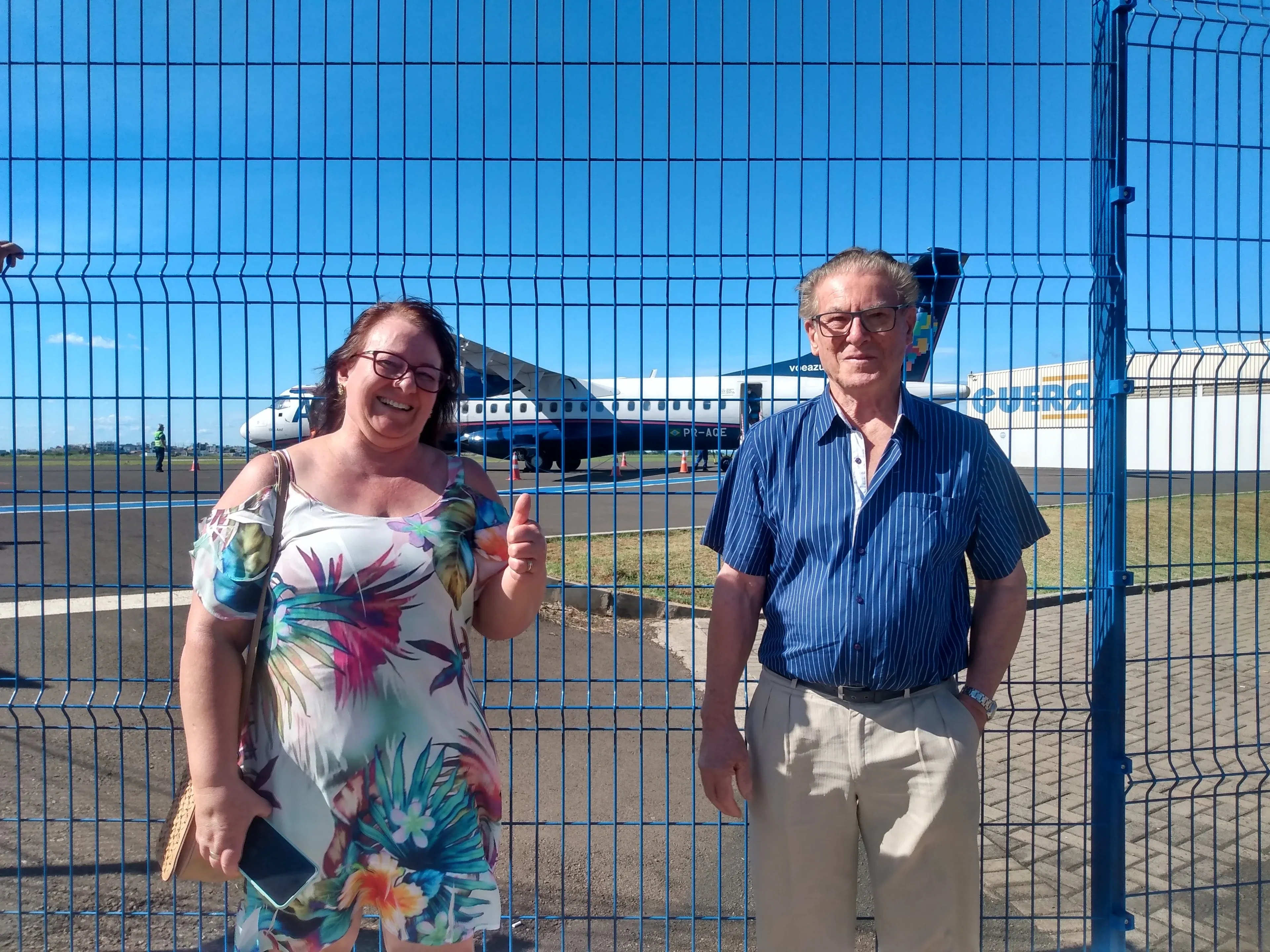 Duas pessoas sorrindo para a câmera em um aeroporto, com um avião visível ao fundo através de uma cerca azul.