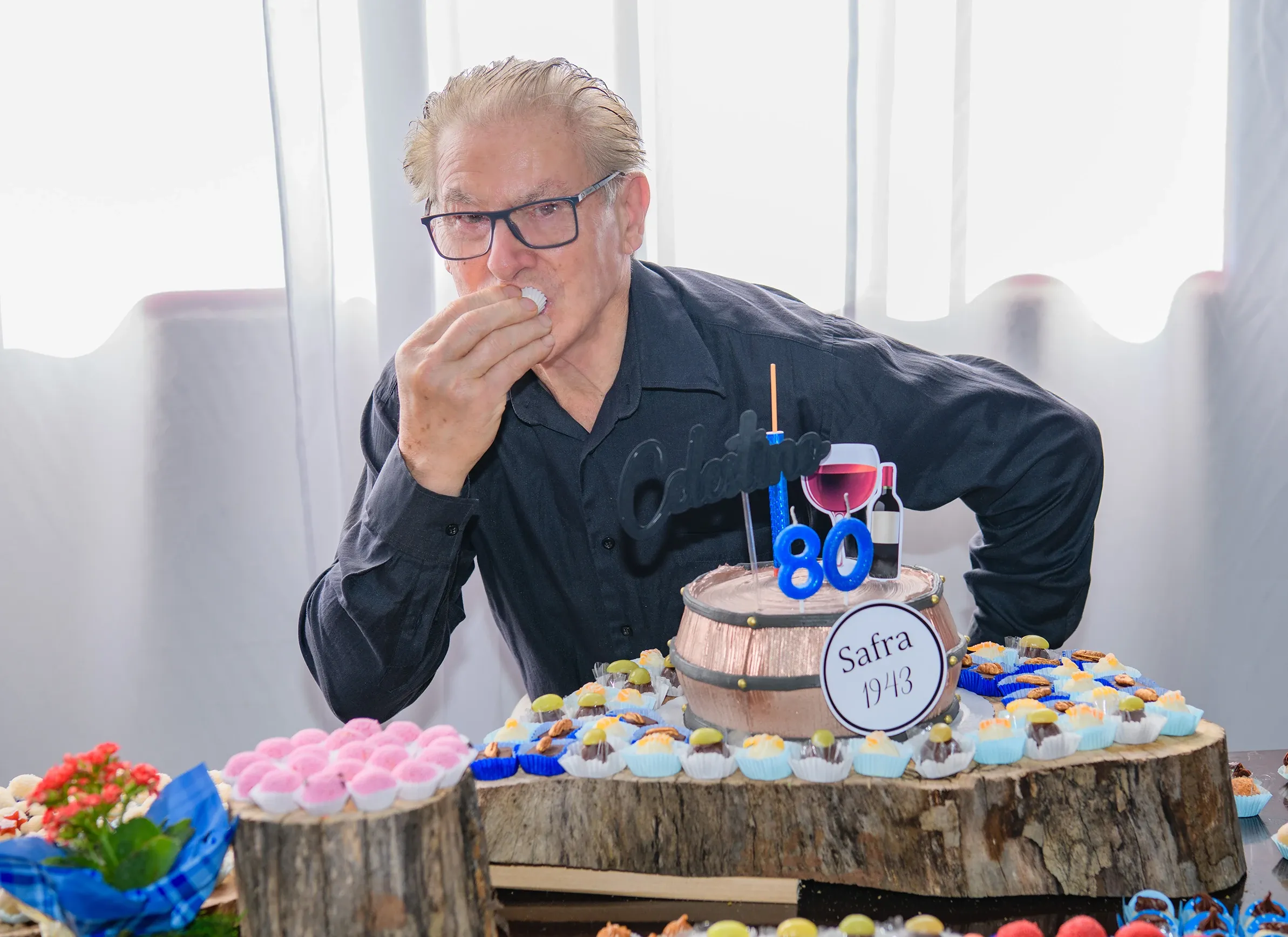 Homem idoso de óculos, vestindo camisa preta, saboreando um doce com a mão direita, atrás de uma mesa de madeira decorada com um bolo de aniversário em formato de barril de vinho e vários doces, em um ambiente interno iluminado por uma janela ao fundo.