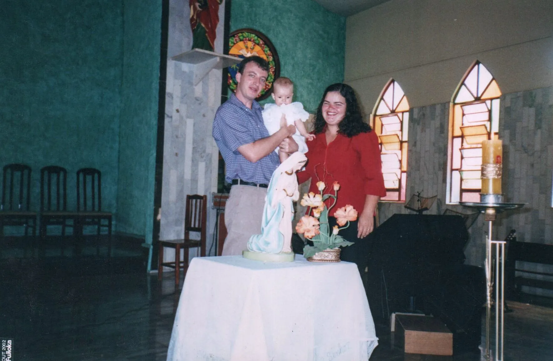 Um homem e uma mulher sorridentes, ambos segurando um bebê vestido de branco, posam para uma foto em um quarto com paredes verdes e azuis, paredes de azulejos e grandes janelas góticas, enquanto se inclinam sobre uma mesa branca com uma estátua da Virgem Maria e um vaso de flores.