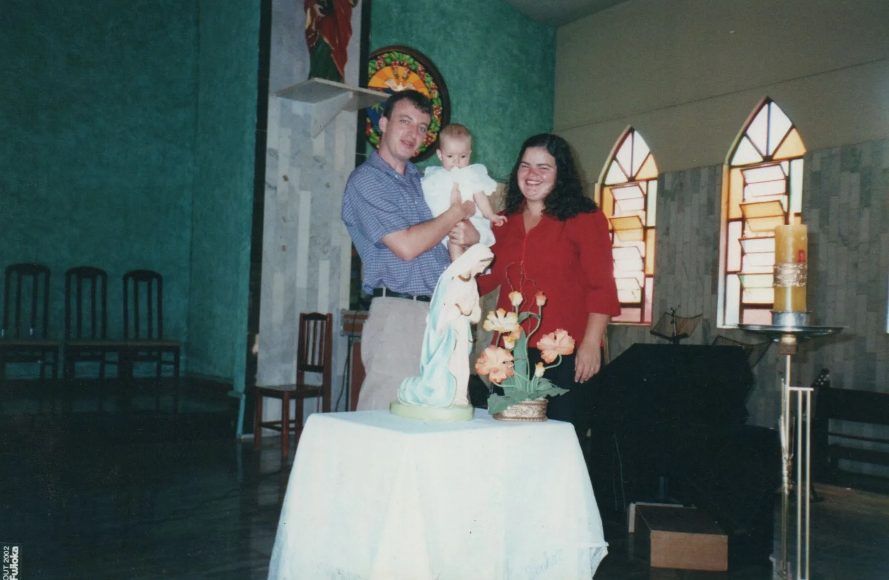 Um homem e uma mulher sorridentes, ambos segurando um bebê vestido de branco, posam para uma foto em um quarto com paredes verdes e azuis, paredes de azulejos e grandes janelas góticas, enquanto se inclinam sobre uma mesa branca com uma estátua da Virgem Maria e um vaso de flores. (Original)