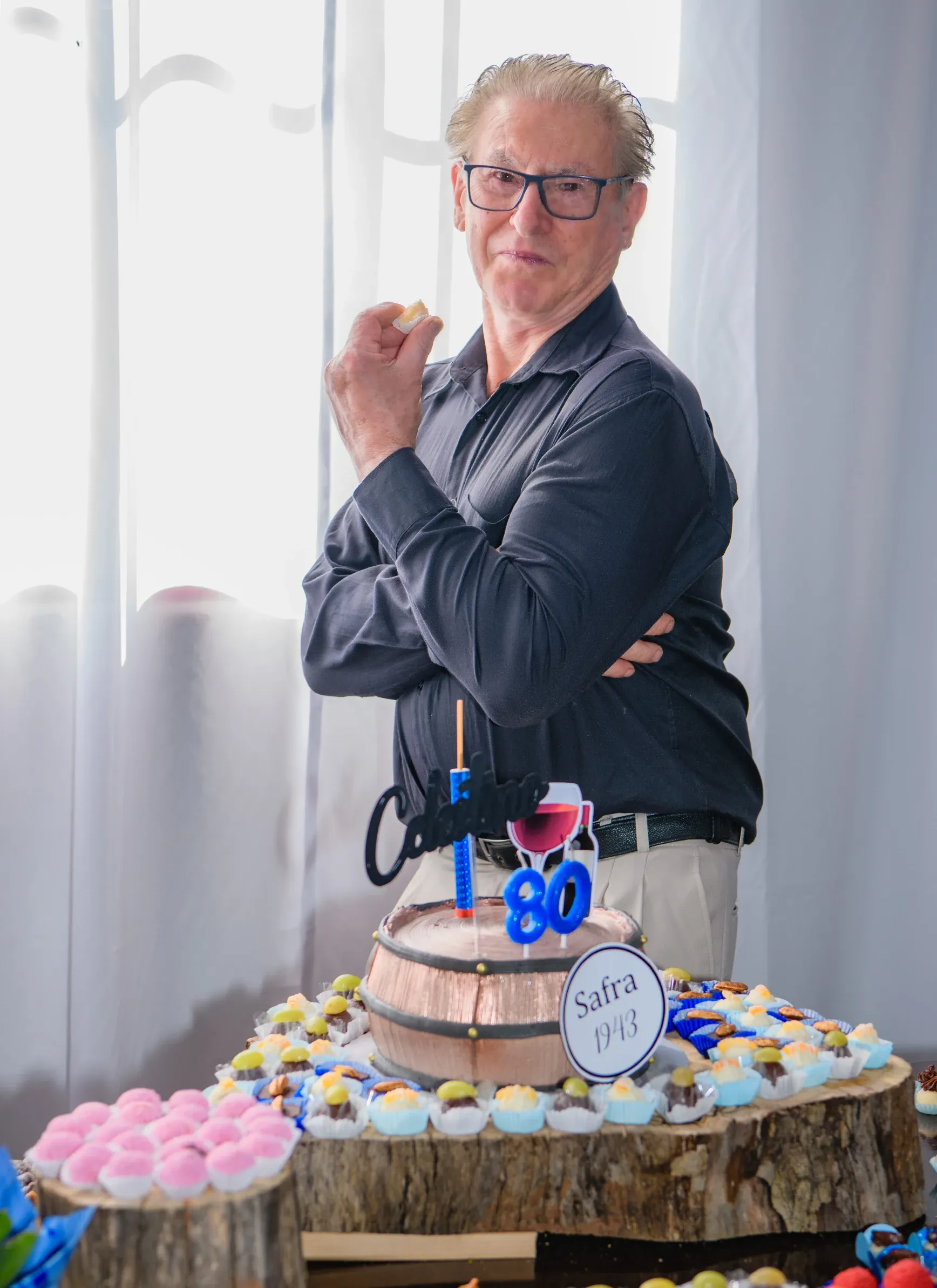 Um homem de óculos e camisa preta, com os braços cruzados, está ao lado de uma mesa com um bolo em formato de barril e doces, com uma janela iluminada ao fundo.