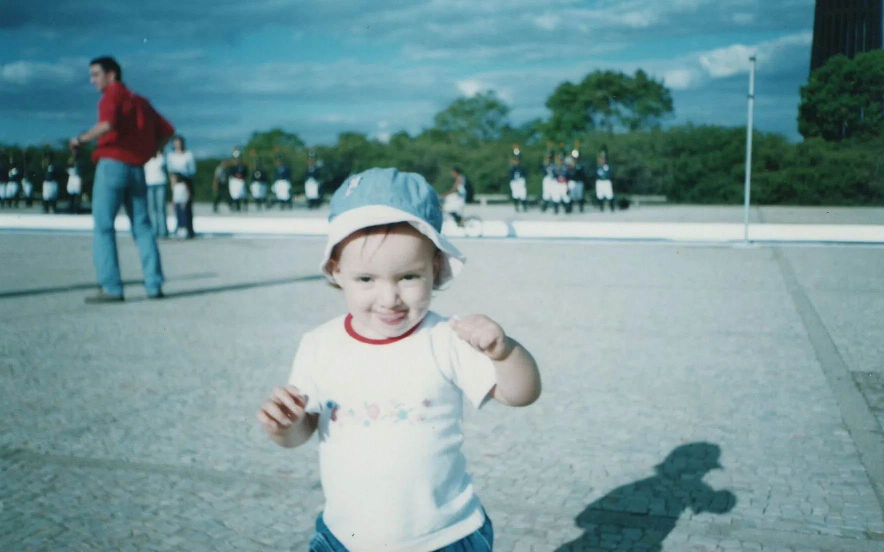 Bebê usando chapéu azul e camiseta branca, sorrindo e estendendo a mão em direção à câmera, com um homem em pé e uma fileira de pessoas ao fundo sob um céu azul. (Original)