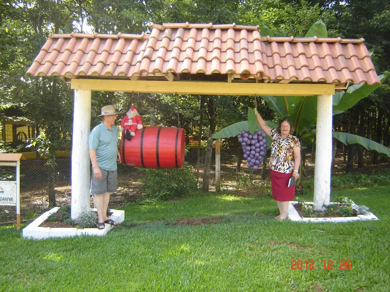 Duas pessoas, uma mulher e um homem, em pé sob uma estrutura com telhado de telhas, com um barril vermelho e um cacho de uvas roxas pendurados, em um gramado verde cercado por árvores.