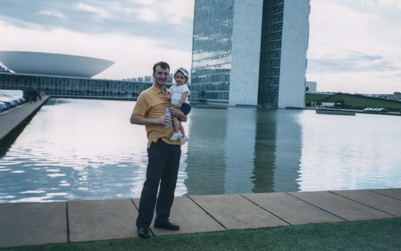 Um homem sorridente de camisa amarela e calças escuras segura um bebê, que usa um chapéu azul e branco, em frente a um grande lago e edifícios modernos de Brasília.