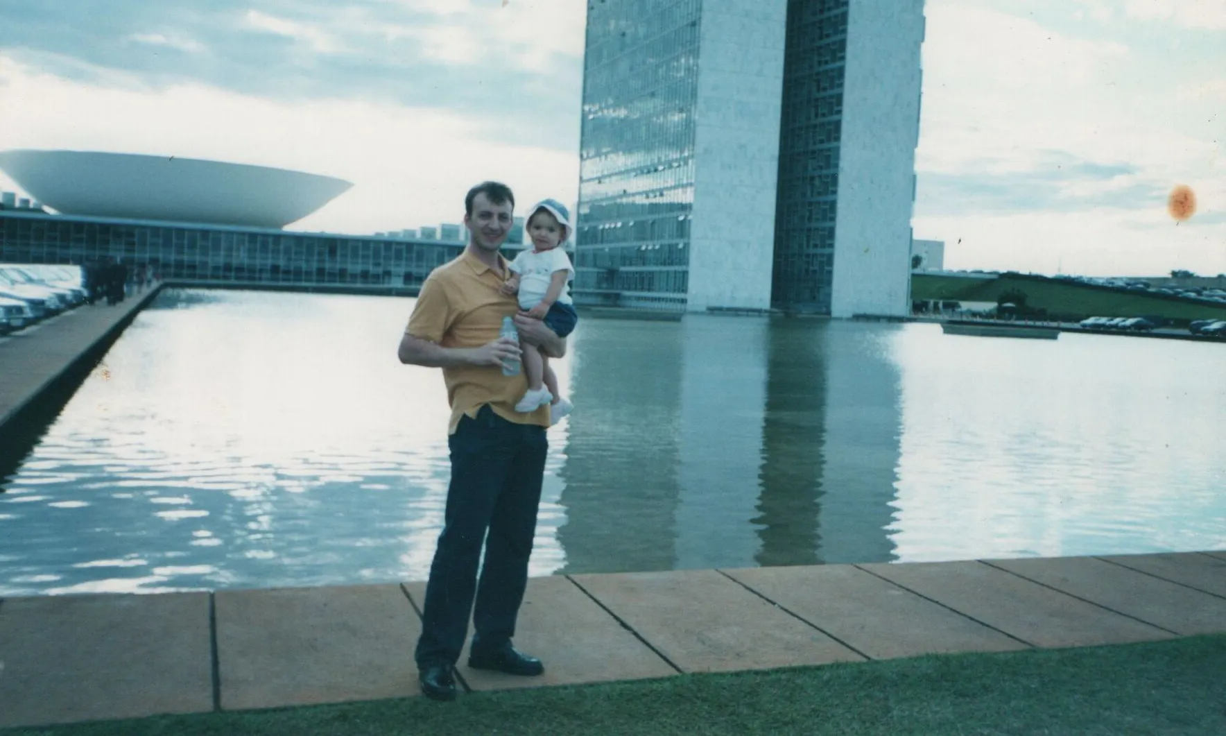 Um homem sorridente de camisa amarela e calças escuras segura um bebê, que usa um chapéu azul e branco, em frente a um grande lago e edifícios modernos de Brasília. (Original)