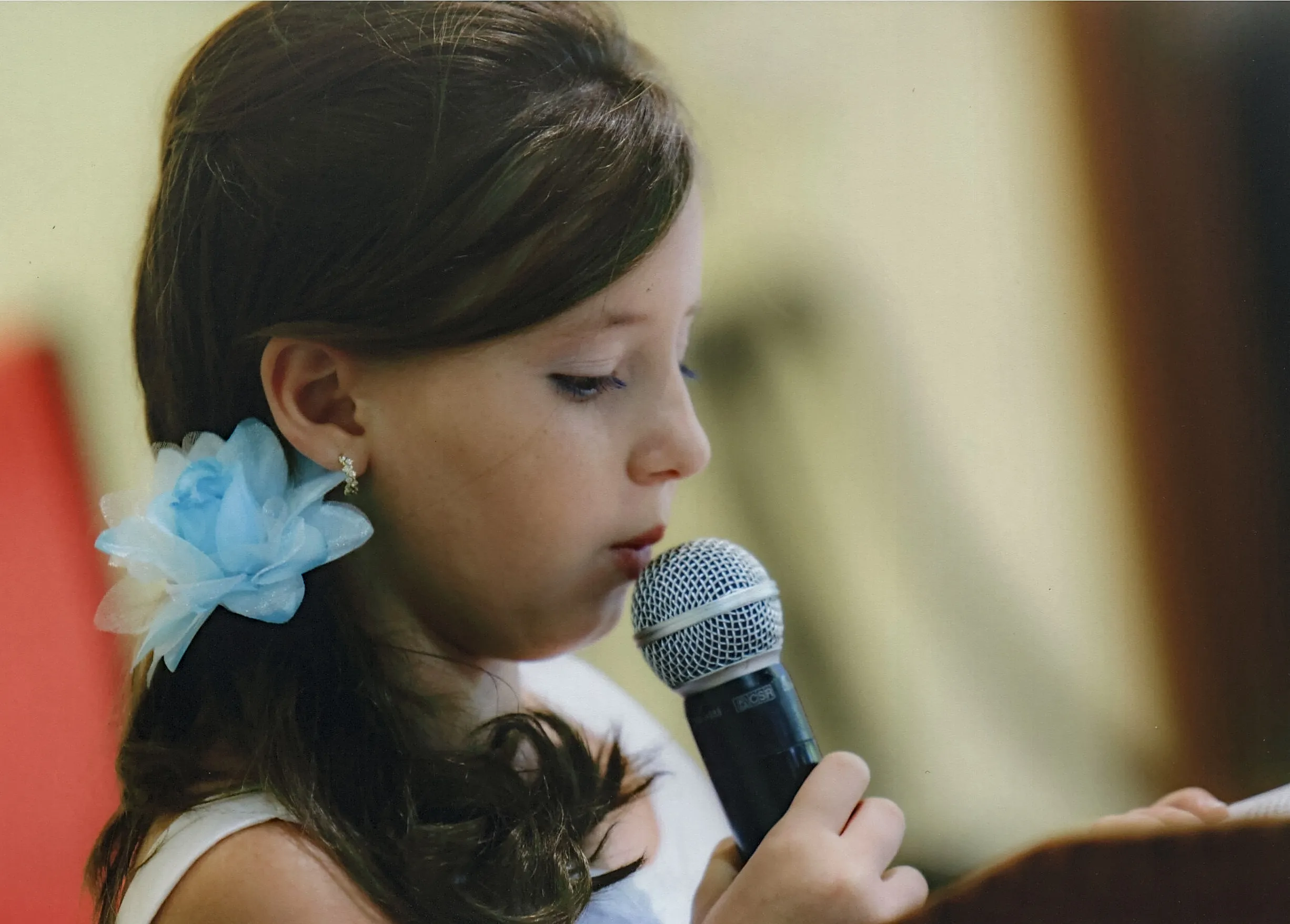 Menina jovem com uma flor azul no cabelo, cantando em um microfone.