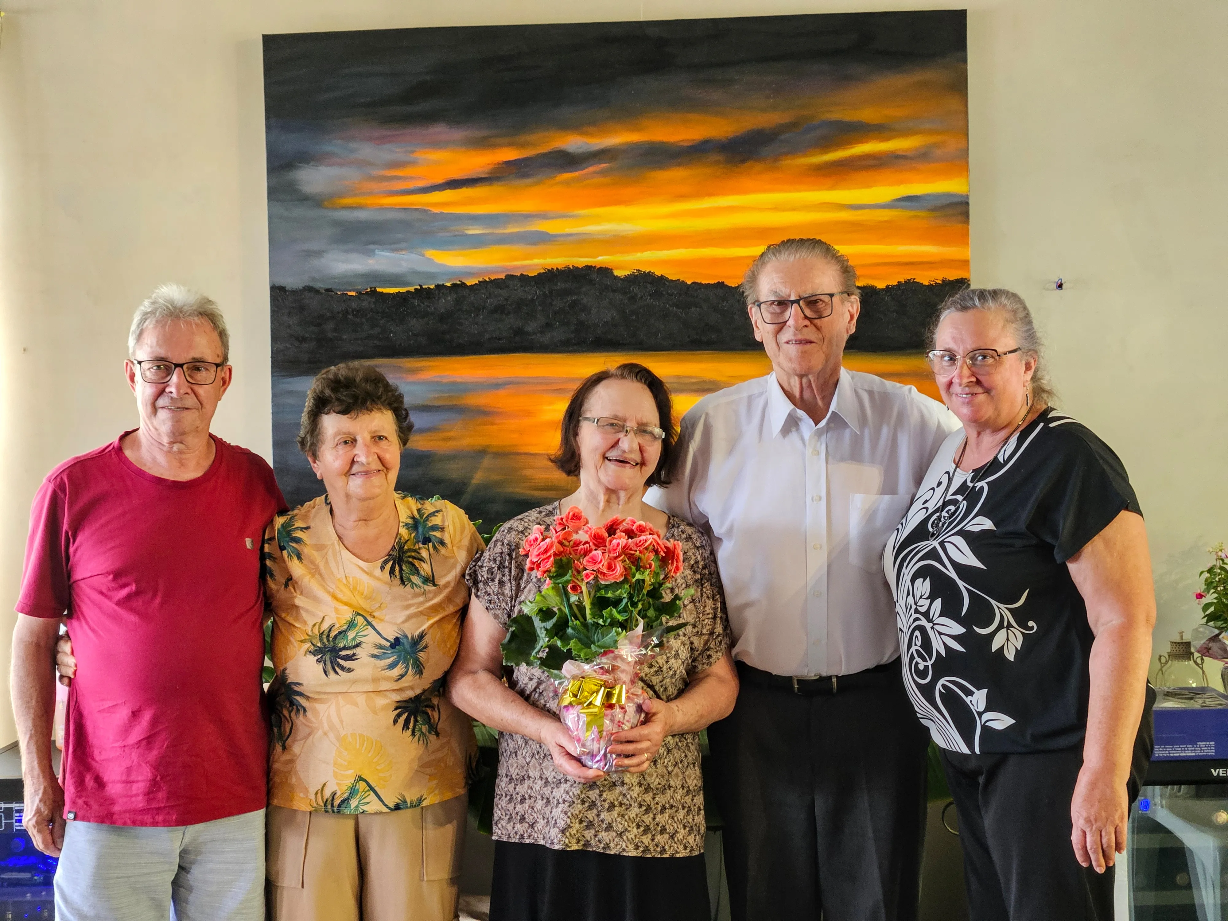 Cinco pessoas posam em frente a uma pintura abstrata. A mulher no meio está segurando um buquê de flores vermelhas.