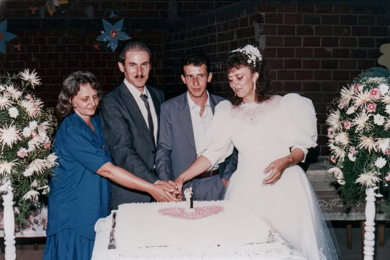 Quatro adultos em volta de um bolo de casamento de três camadas, com a noiva de branco e o noivo de terno cinza ao lado dela, enquanto uma mulher de azul escuro e um homem de terno preto os ajudam a cortar o bolo.