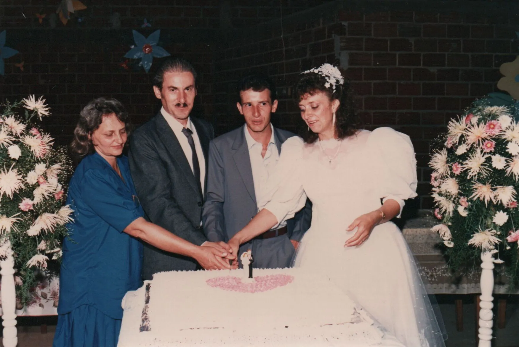 Quatro adultos em volta de um bolo de casamento de três camadas, com a noiva de branco e o noivo de terno cinza ao lado dela, enquanto uma mulher de azul escuro e um homem de terno preto os ajudam a cortar o bolo. (Original)
