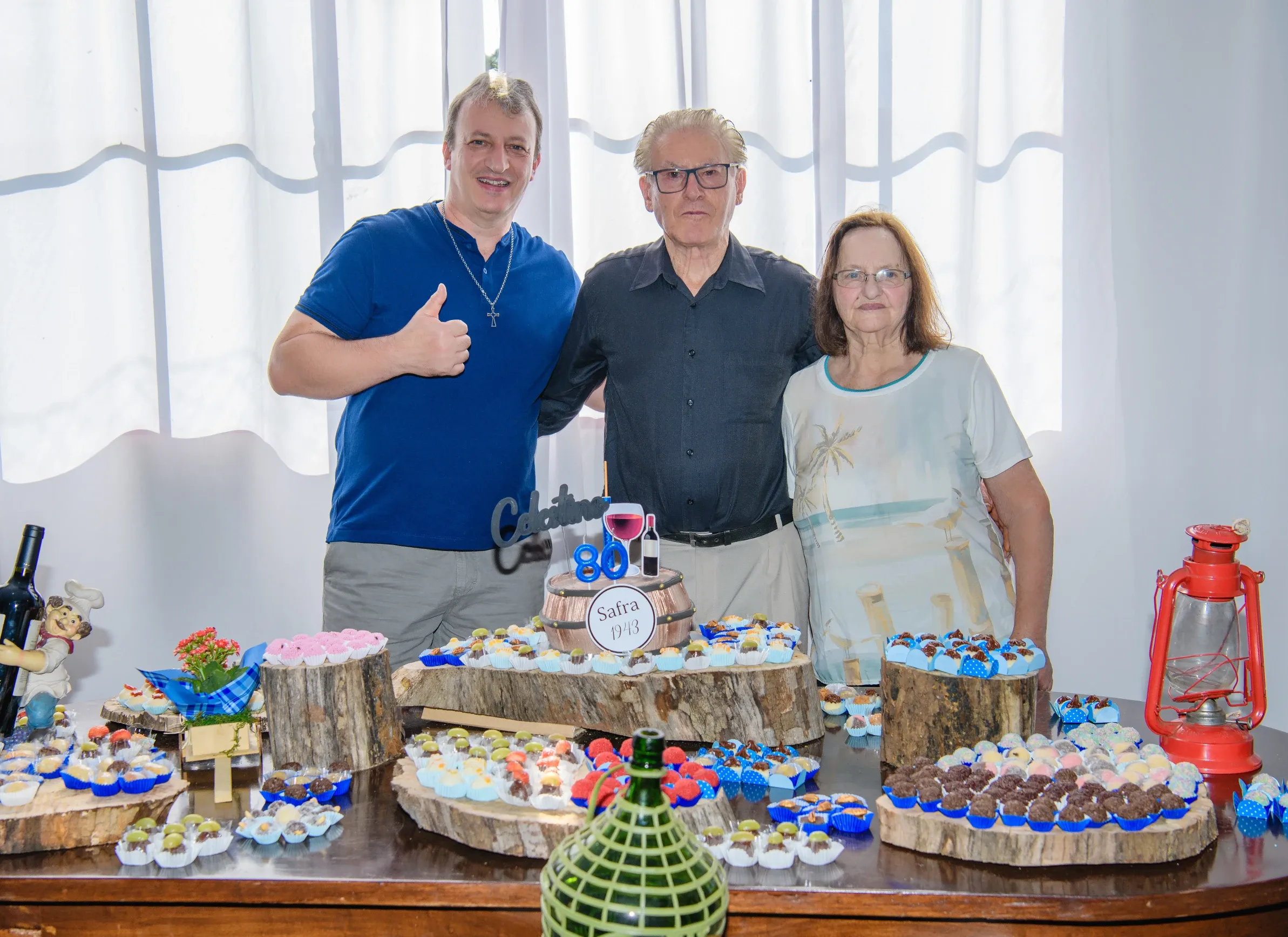 Um homem de camisa azul, um homem idoso de óculos e camisa preta e uma mulher de blusa branca e óculos posam atrás de uma mesa com doces e um bolo de 80º aniversário.