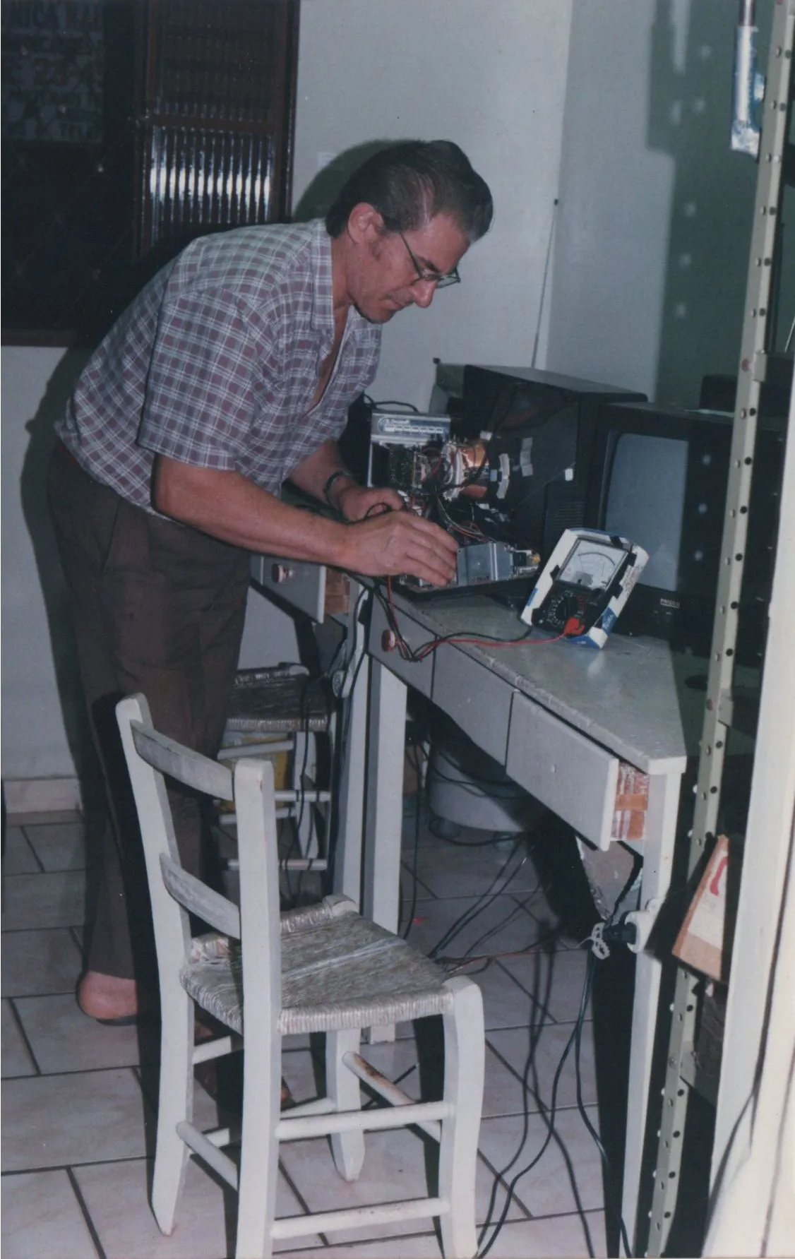Um homem de meia-idade com óculos, camisa xadrez e calças marrons está inclinado sobre uma mesa de trabalho cheia de equipamentos eletrônicos, incluindo um multímetro, enquanto conserta uma televisão antiga em uma sala com pouca iluminação. (Original)