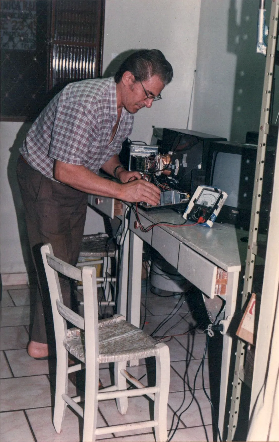 Um homem de meia-idade com óculos, camisa xadrez e calças marrons está inclinado sobre uma mesa de trabalho cheia de equipamentos eletrônicos, incluindo um multímetro, enquanto conserta uma televisão antiga em uma sala com pouca iluminação.