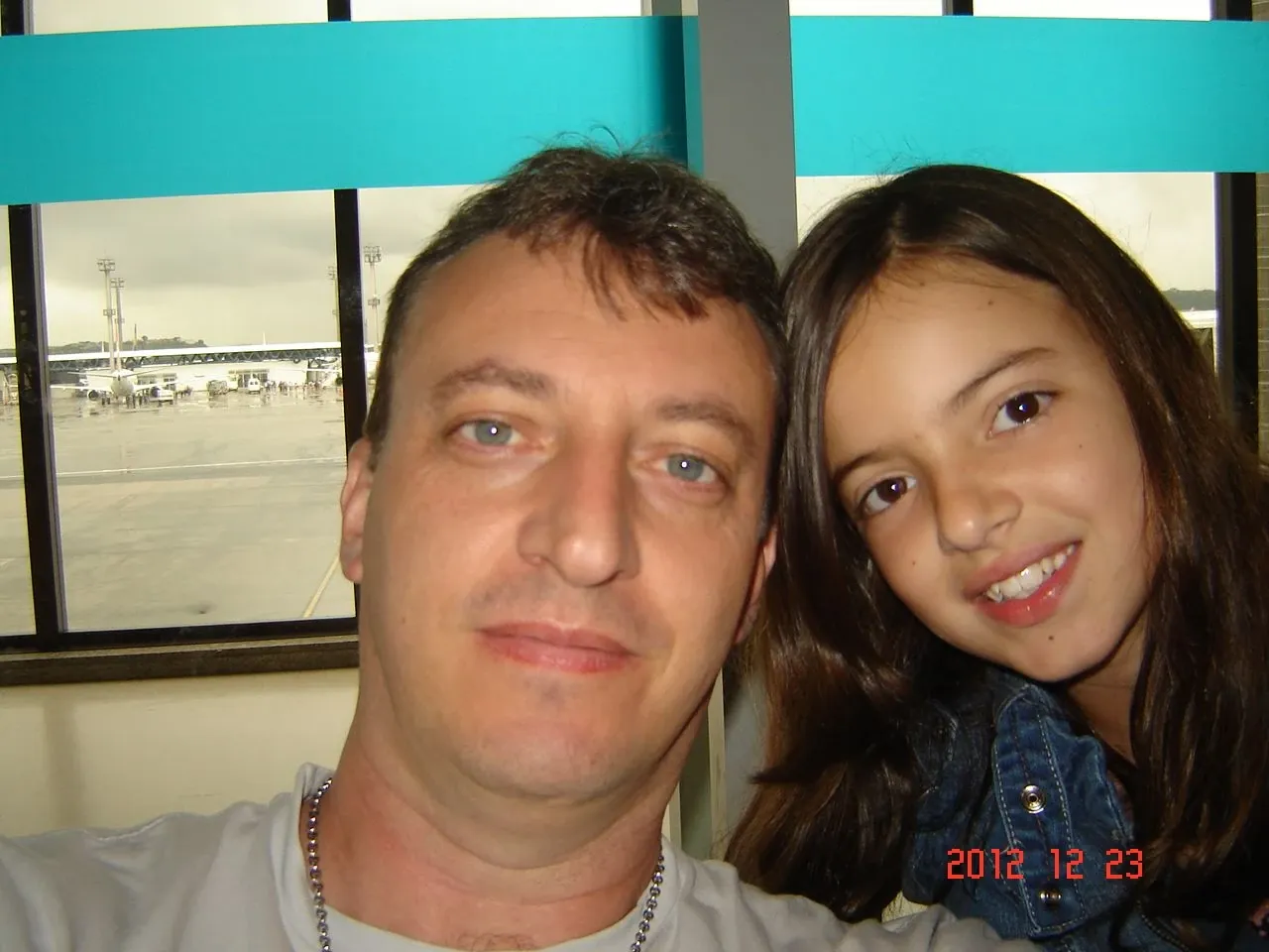 Homem sorridente e criança em primeiro plano, com o aeroporto visível através da janela.