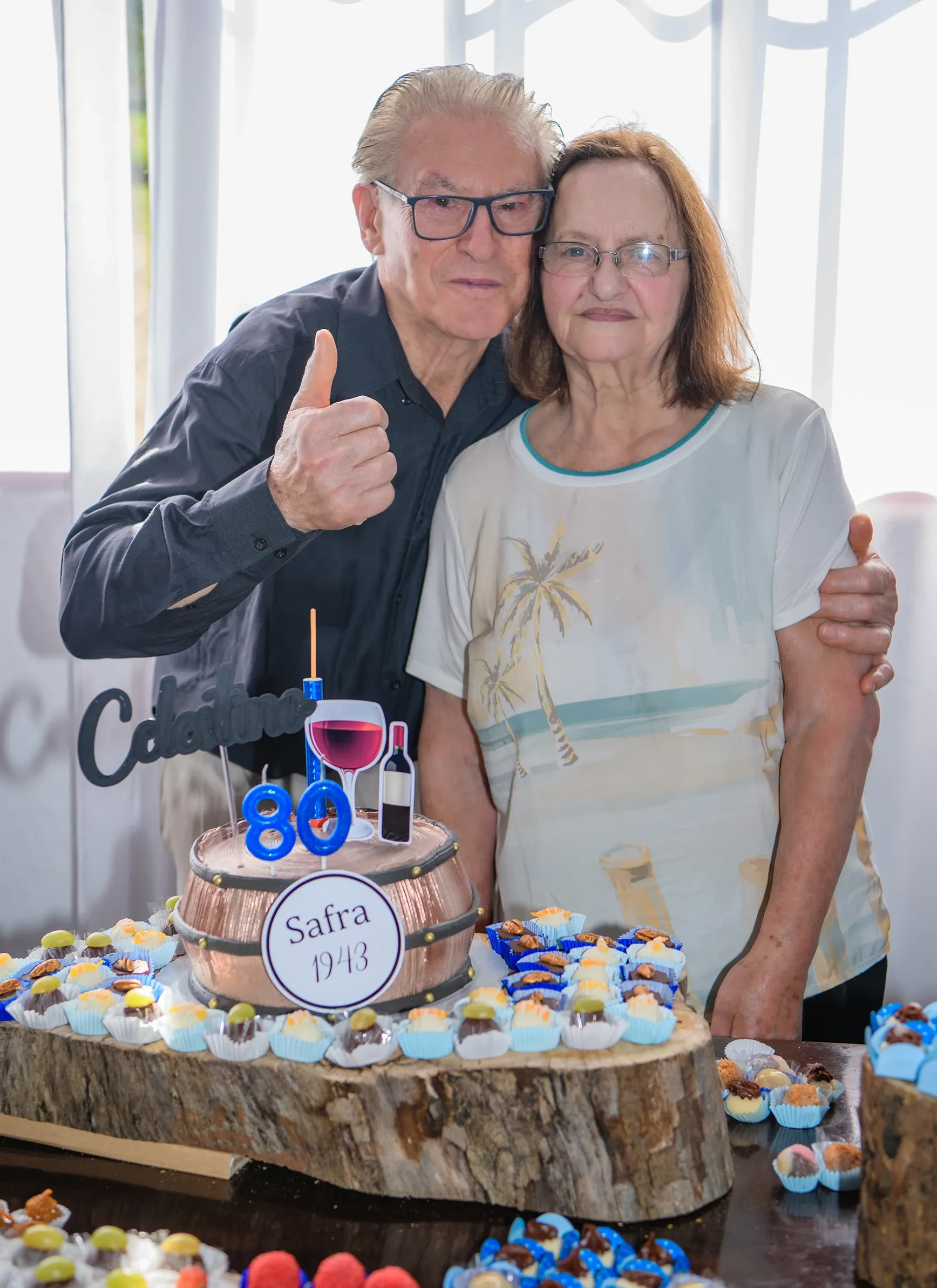 Um homem idoso de óculos e camisa escura abraça uma mulher idosa de óculos e blusa branca estampada, ambos sorrindo e olhando para a frente, atrás de um bolo de aniversário de barril de vinho e doces em uma mesa de festa.