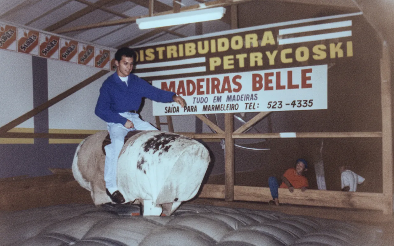 Um homem de camisa azul e calças jeans está montado em um touro mecânico em um evento interno. Um banner com o texto "DISTRIBUIDORA PETRYKOSKI MADEIRAS BELLE" aparece atrás dele.